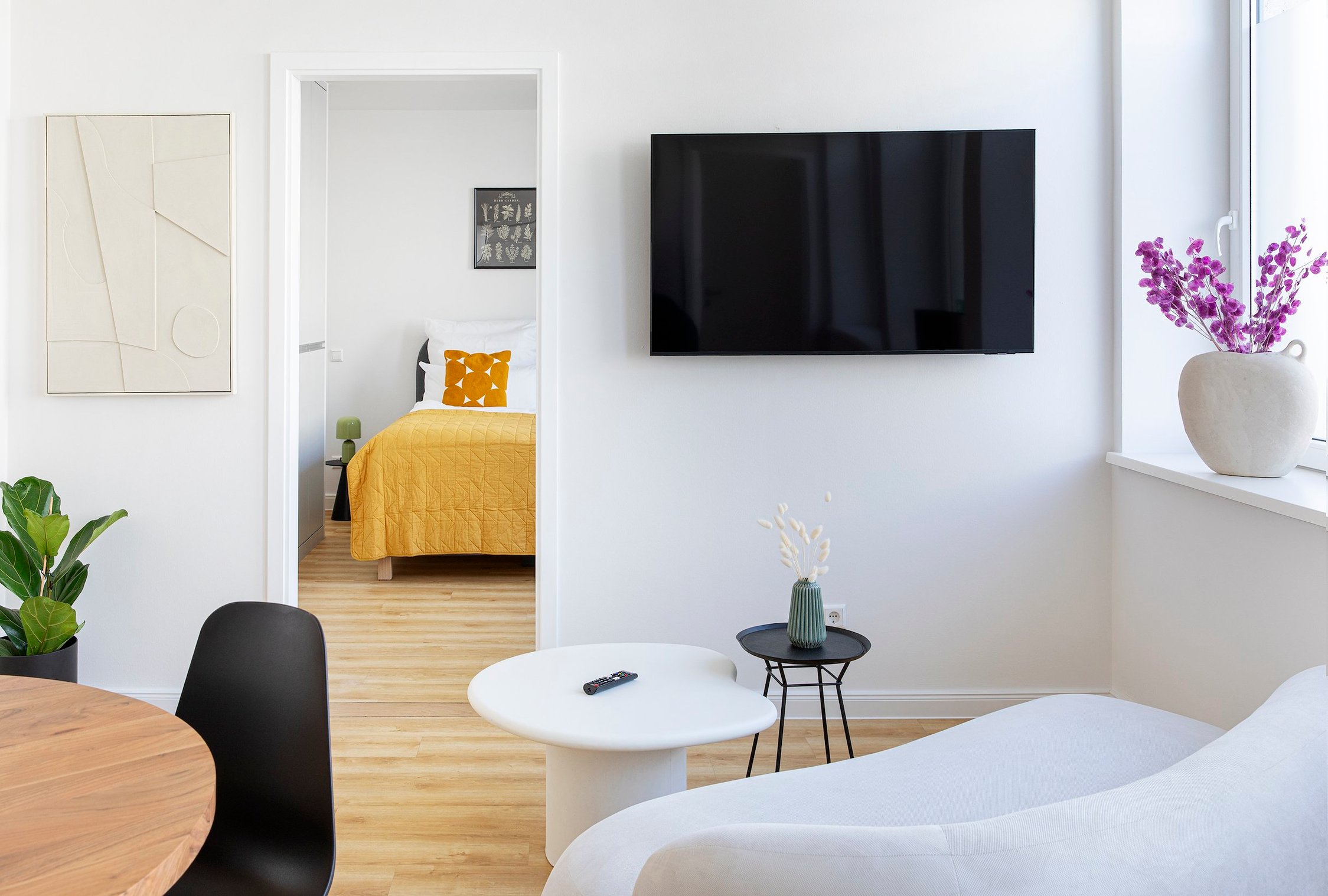 Heller Wohnbereich mit TV, Sofa, Holztisch, offener Tür zu Schlafzimmer mit gelber Decke