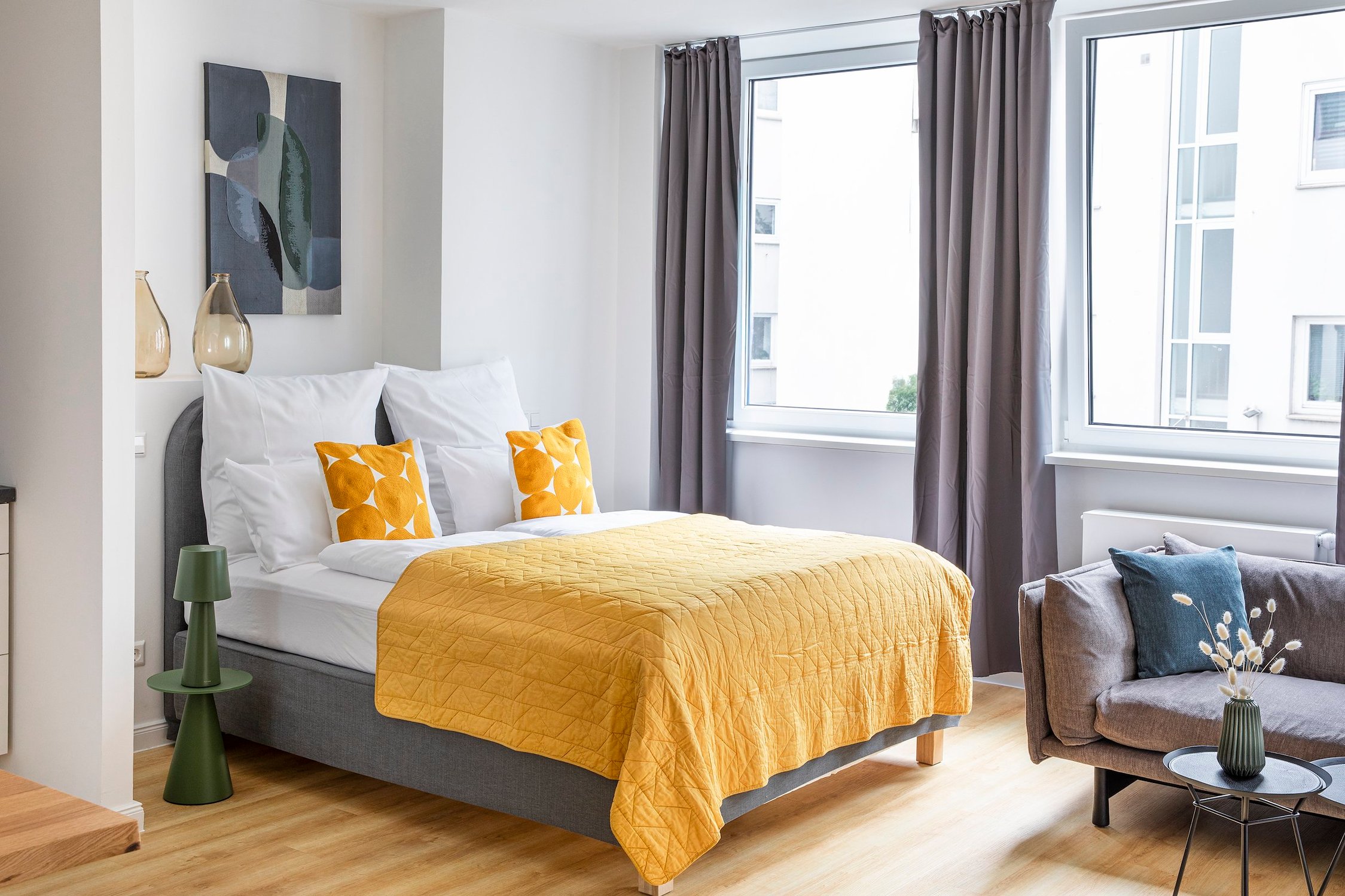Schlafzimmer mit grauem Bett, gelber Tagesdecke, dahinter zwei Fenster und Couch