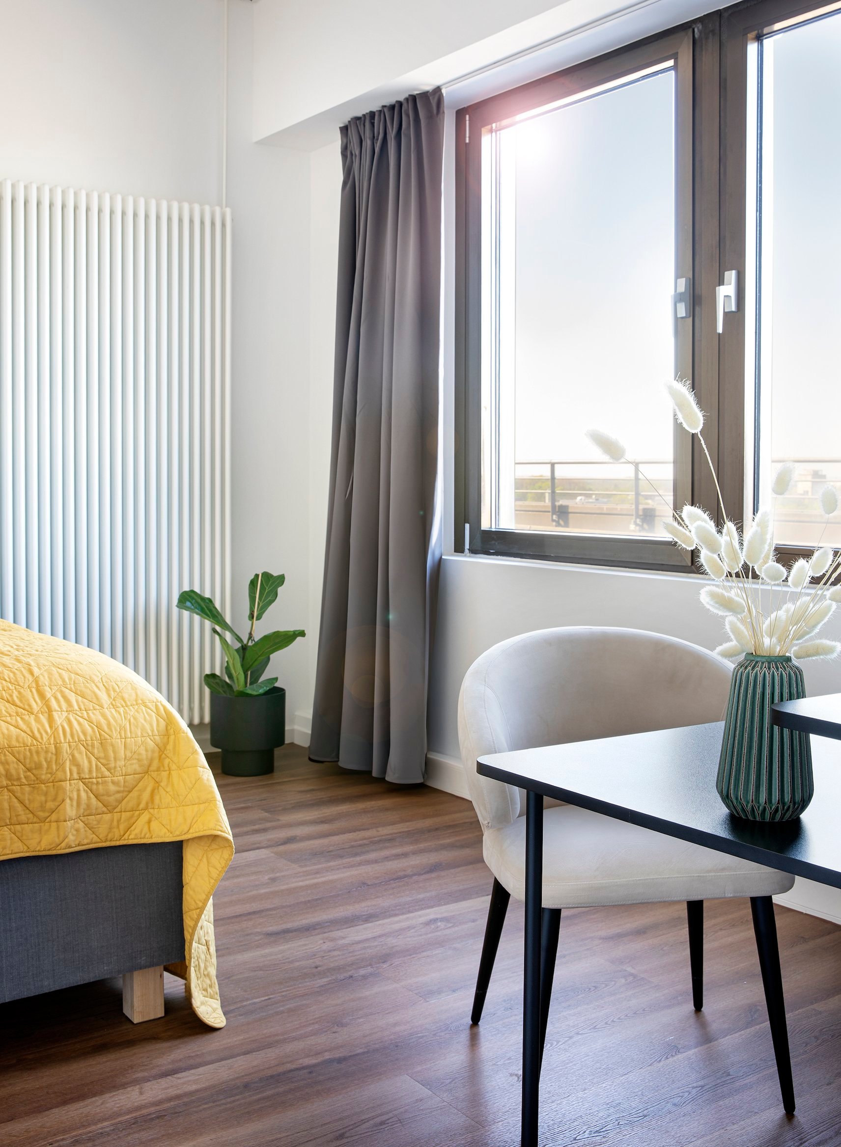 Schlafzimmer mit Balkonblick, hölzernem Boden, Heizung, Bett, Pflanze, Stuhl und Vase auf Tisch