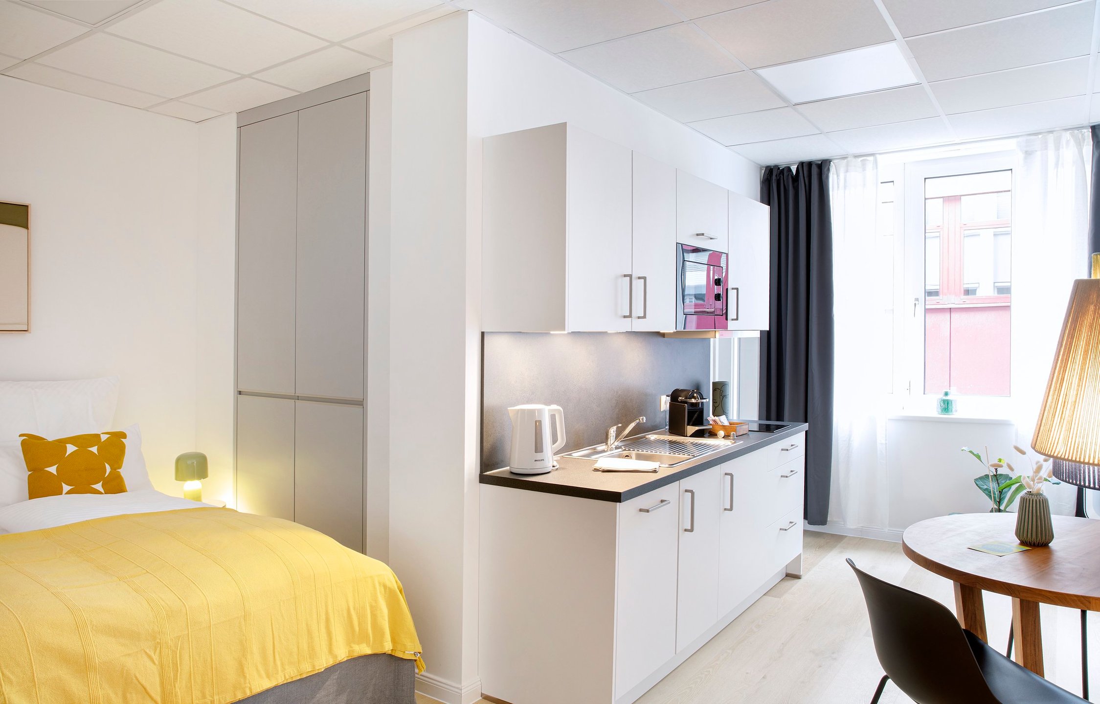 Modernes Studioapartment mit gelb bezogenem Bett, integrierter weißer Küchenzeile und Holztisch