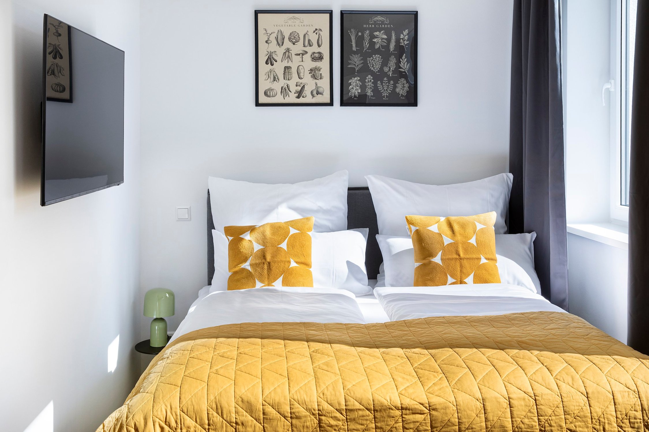 Schlafzimmer mit Doppelbett, senfgelber Tagesdecke, abstrakten Kissen und Wand-TV