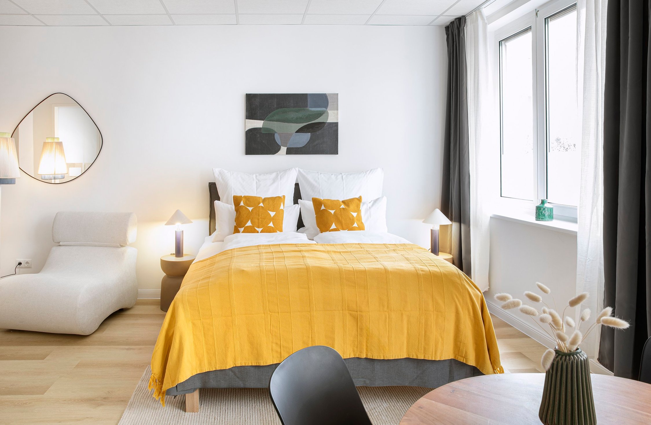 Schlafzimmer mit gelber Tagesdecke, Bett, Liege, Wandspiegel und großem Fenster