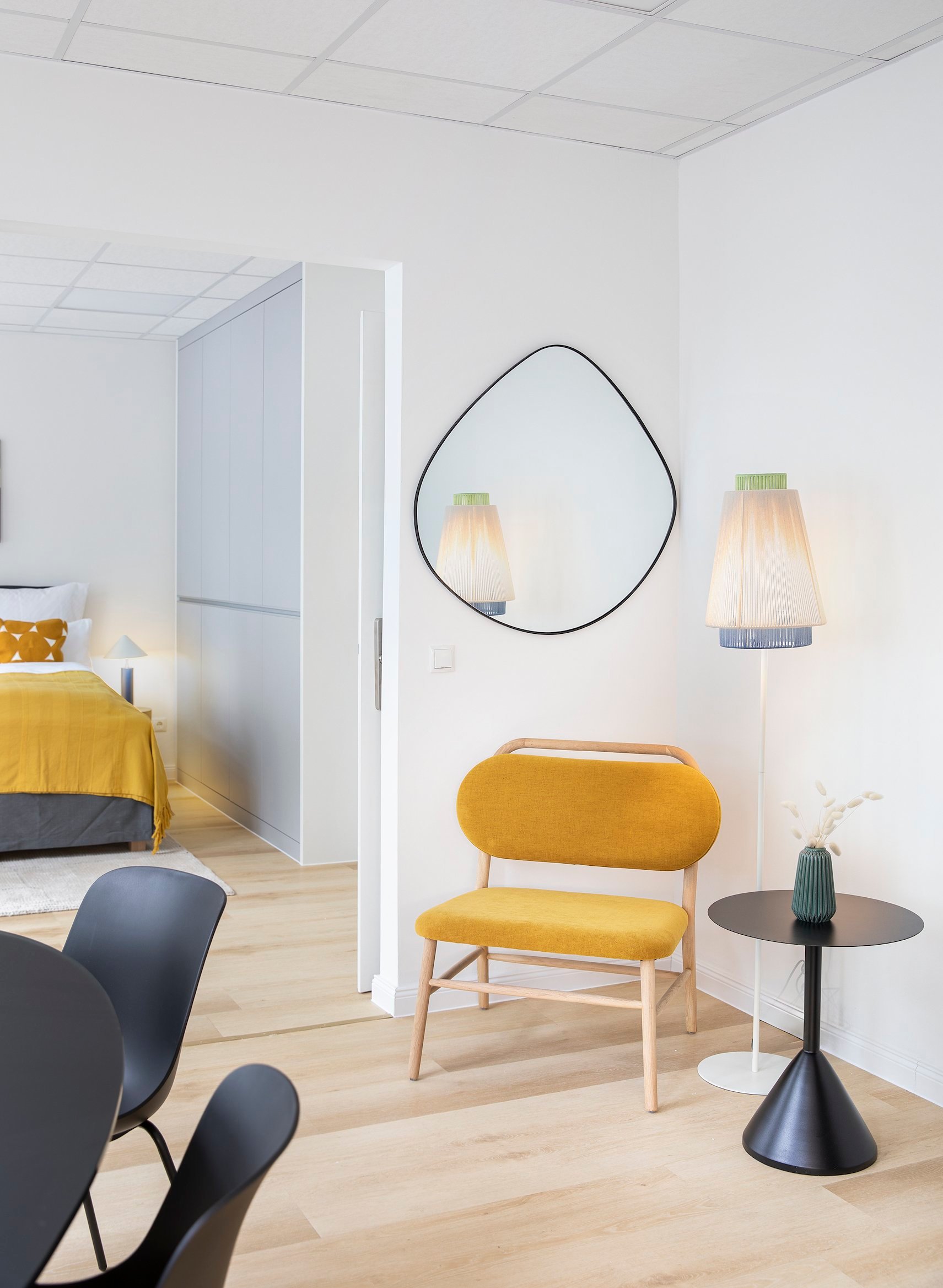 Helles Zimmer mit gelbem Sessel an weißer Wand, ovalem Spiegel und schwarzem Beistelltisch