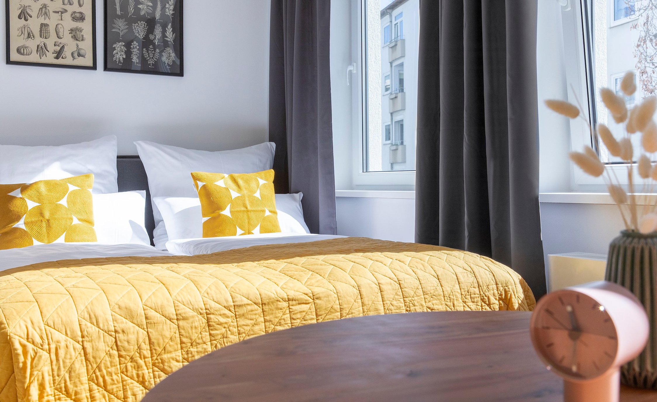 Bett mit gelber Steppdecke und Kissen, helles Fenster mit dunklen Vorhängen, runder Holztisch im Vordergrund