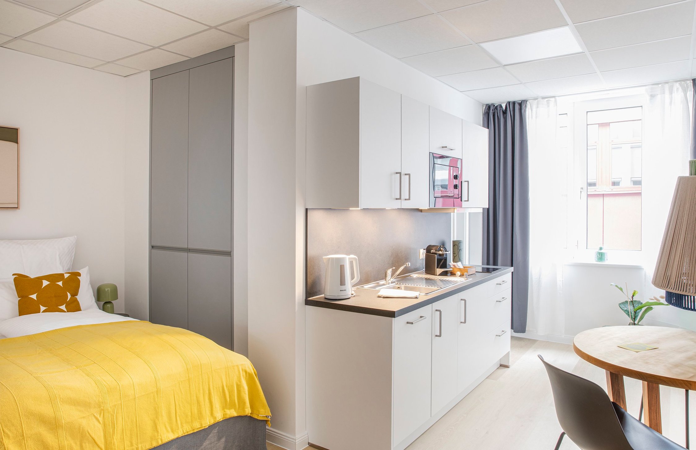 Studio-Apartment mit Bett, grauer Küchenzeile und Holztisch, helle Einrichtung