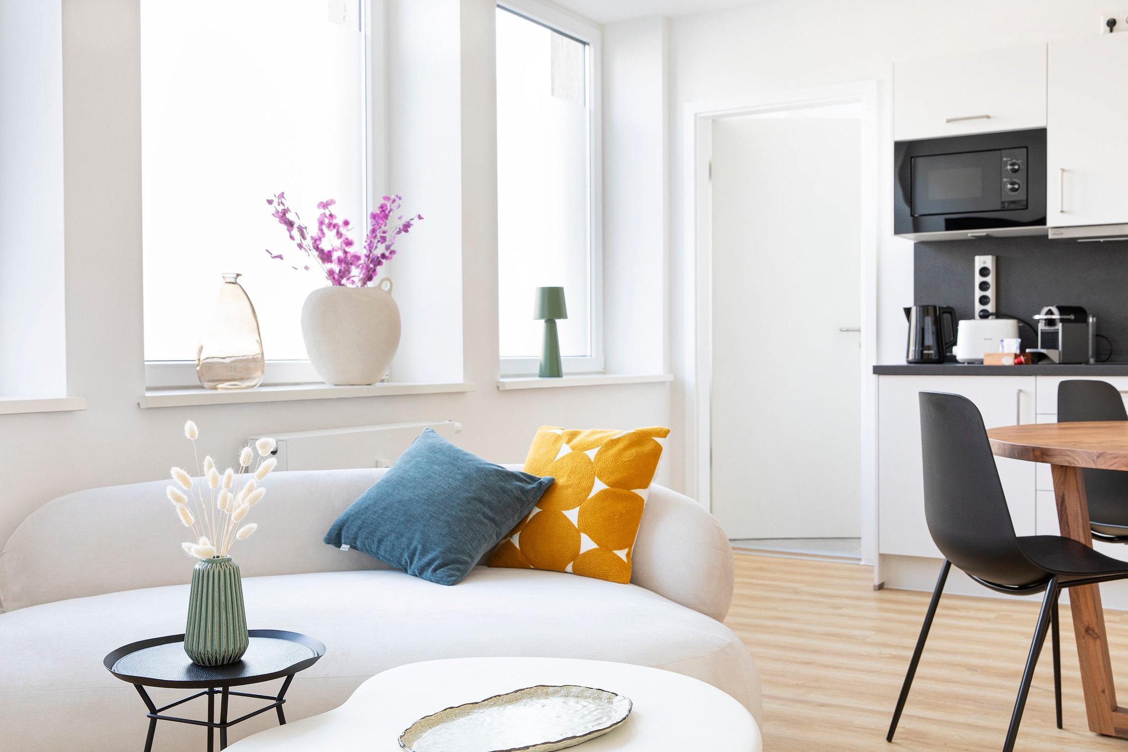 Wohnküche mit weißem Sofa, Holz-Esstisch, schwarzem Hocker und schlichter Dekoration