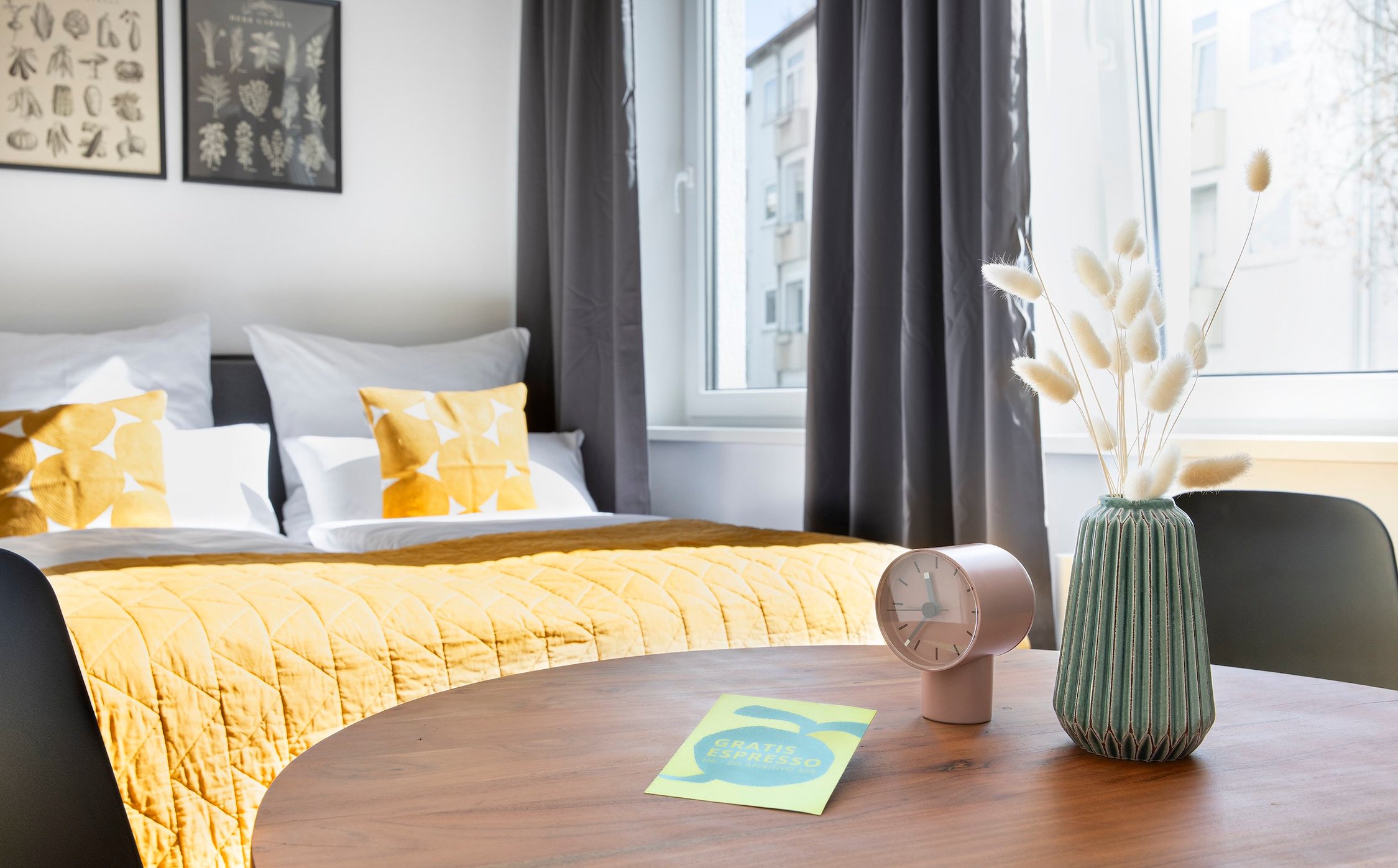 Schlafzimmer mit gelber Tagesdecke, dahinter ein runder Holztisch, darauf Vase und Uhr