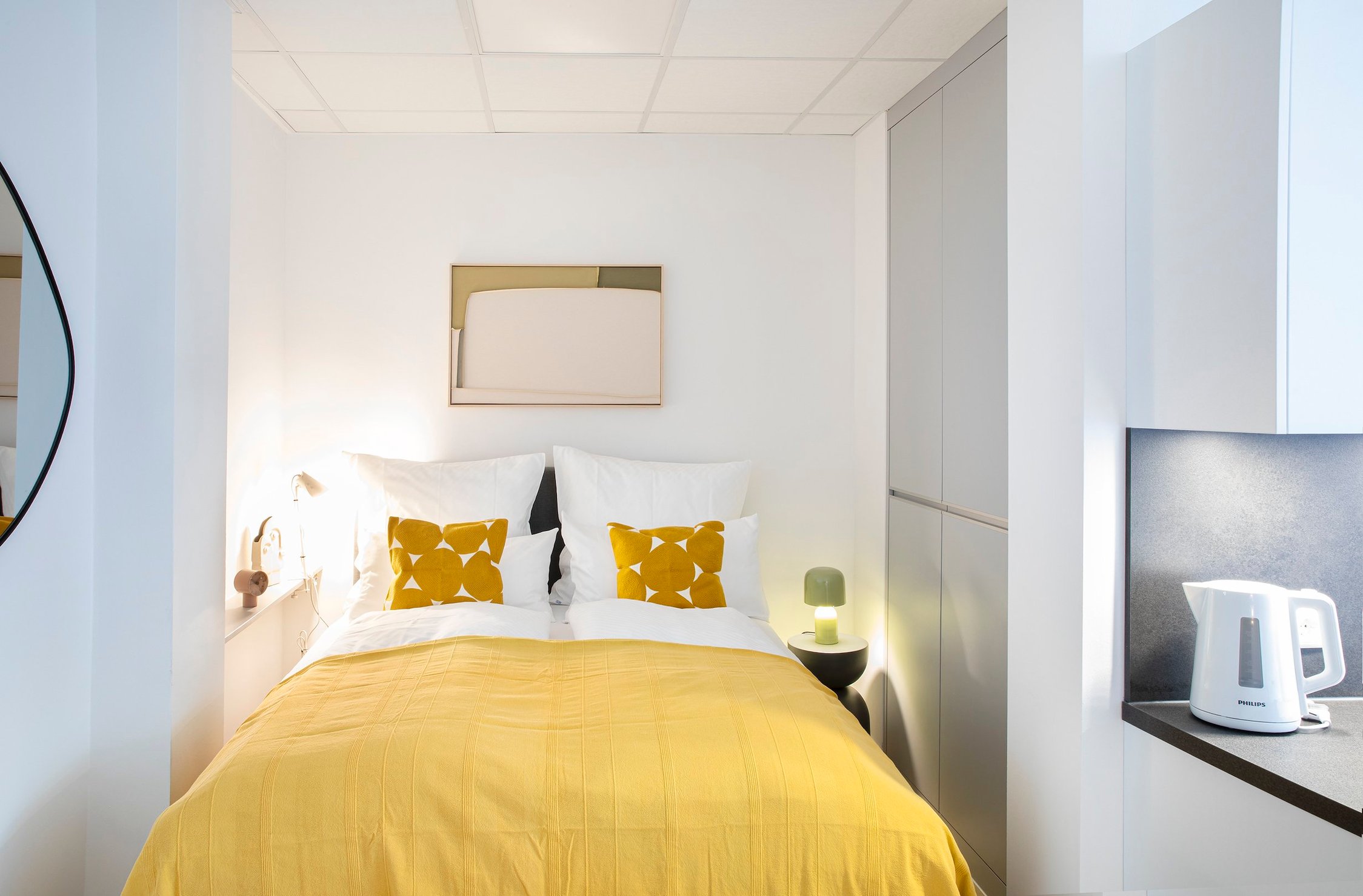 Helles Schlafzimmer mit Einzelbett, gelber Decke, zwei Kissen und weißem Wasserkocher auf Ablage