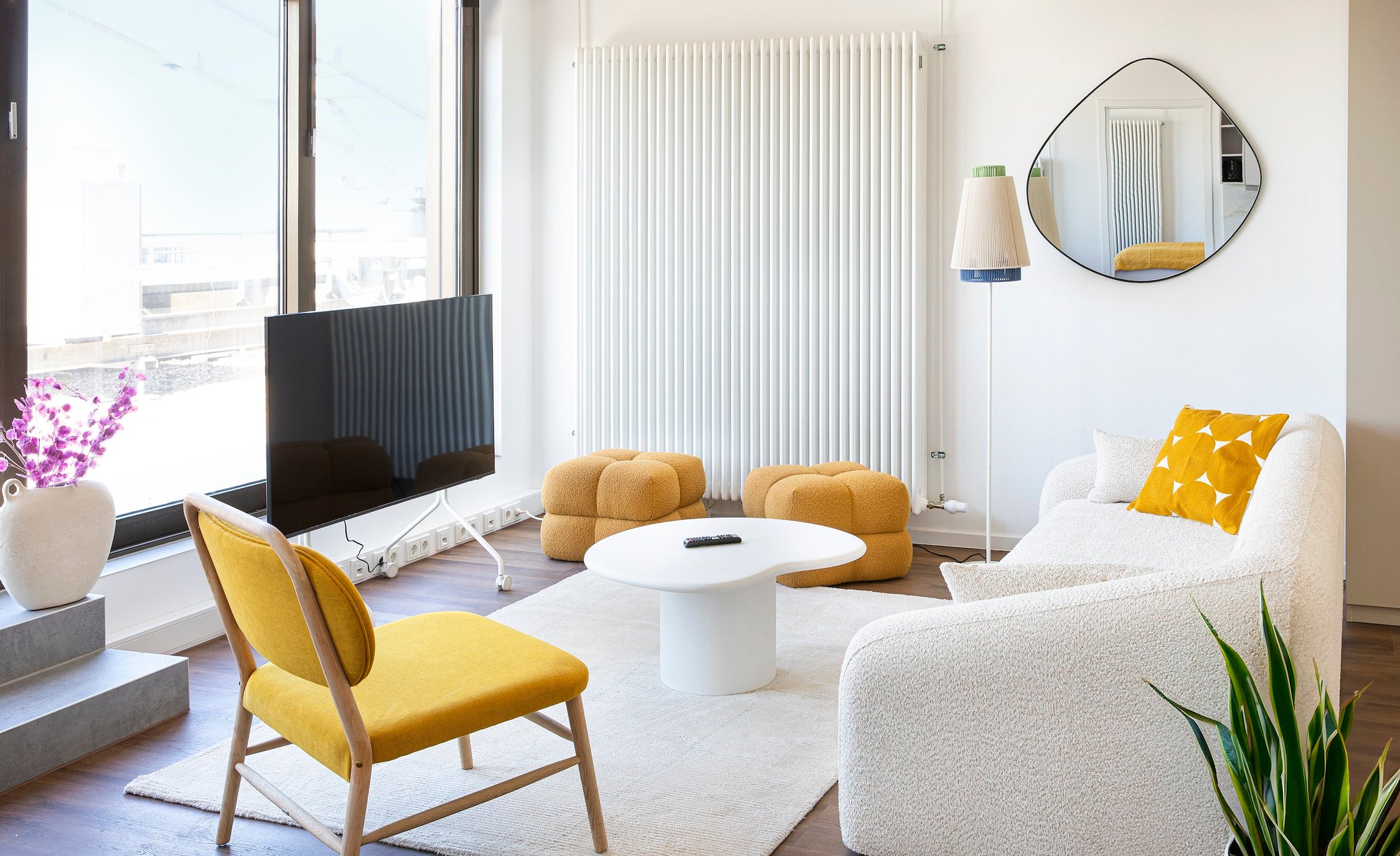 Helles Wohnzimmer mit weißem Sofa, gelbem Sessel, TV und Beistelltisch auf Teppich, weiße Wände