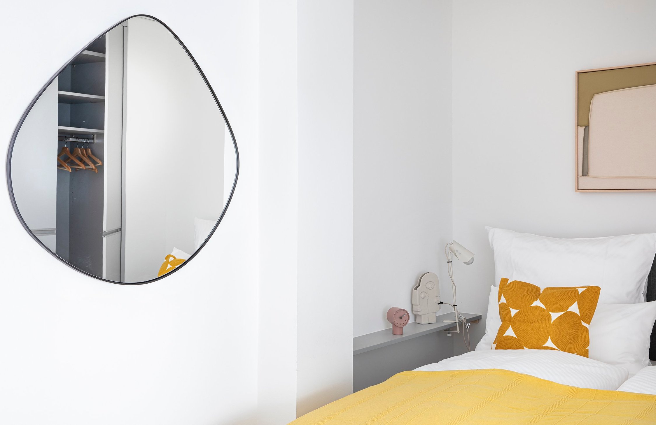Schlafzimmer mit spiegelnder organischer Form, weißer Bettlaken und gelbem Kissen auf dem Bett