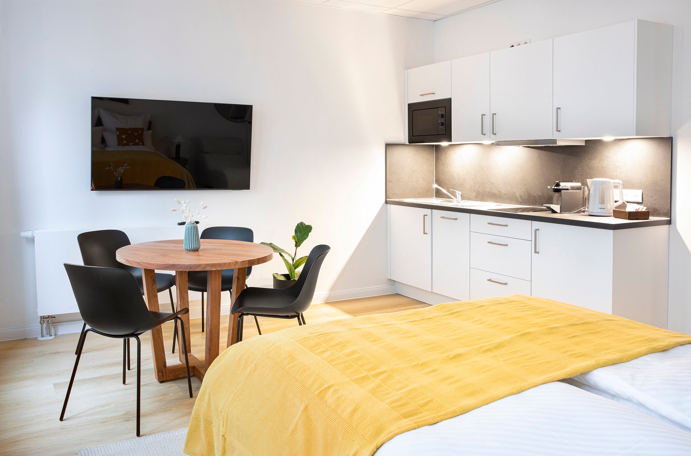 Einraumwohnung mit weißer Küchenzeile, Holztisch, vier schwarzen Stühlen und gelber Bettdecke
