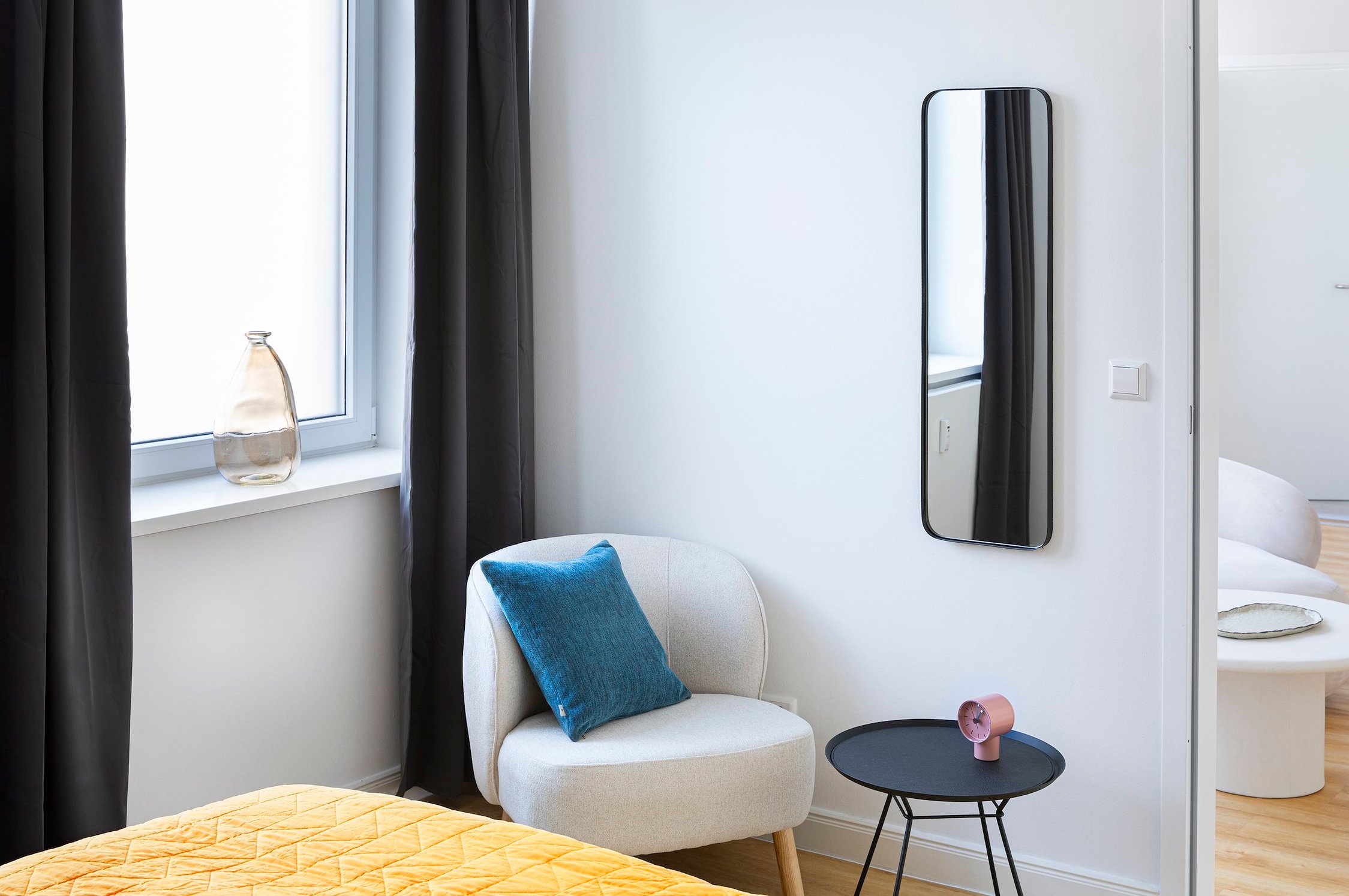 Schlafzimmer mit weißem Stoffsessel, schwarzem Beistelltisch und schlankem Wandspiegel