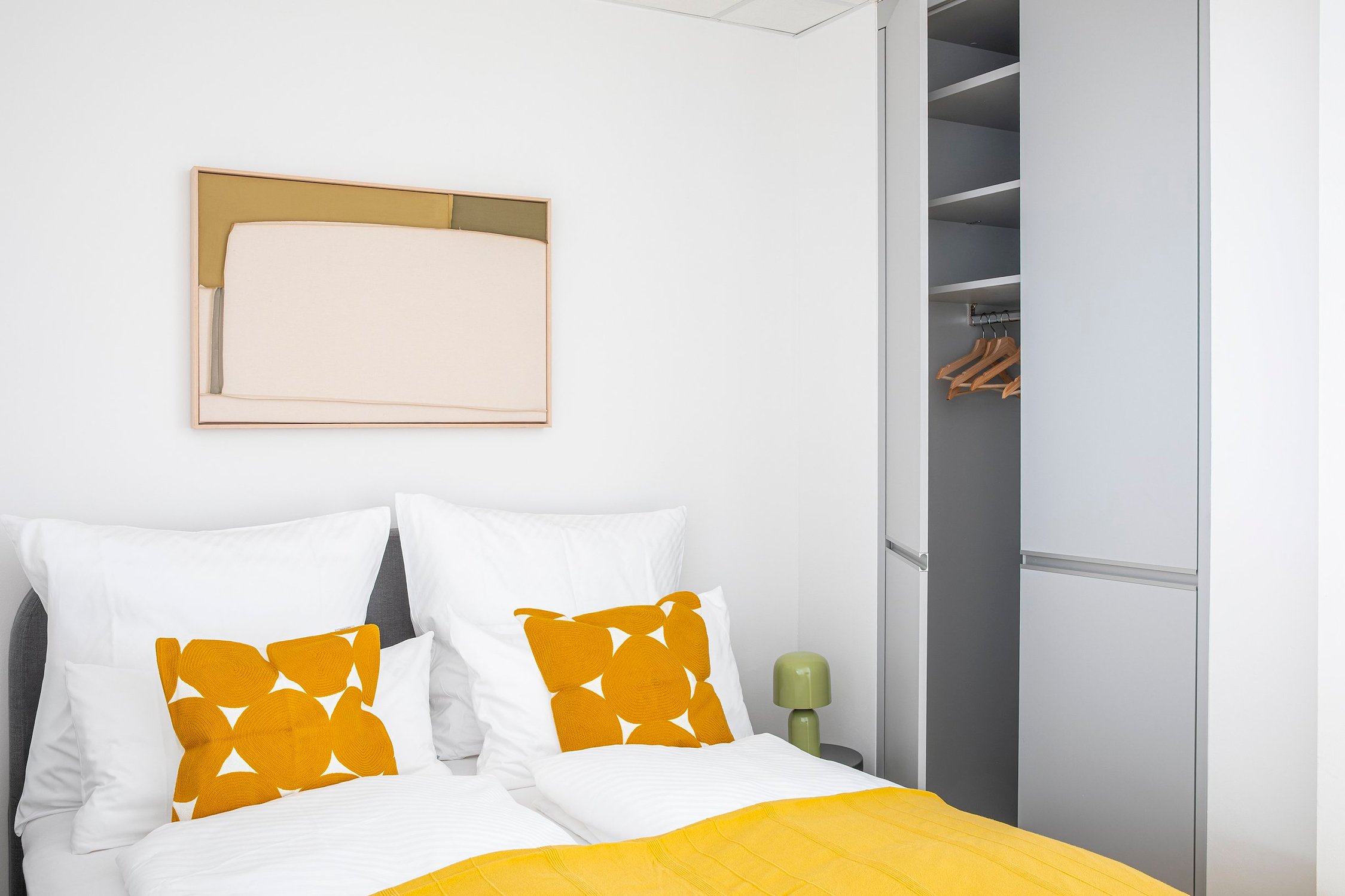 Schlafzimmer mit weißer Bettwäsche, gelben Kissen und geöffnetem grauen Kleiderschrank