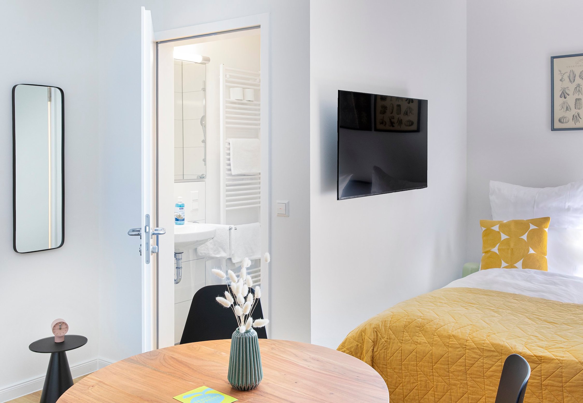 Helles Zimmer mit kleinem Holztisch, Bett mit gelber Decke und offenem Bad dahinter