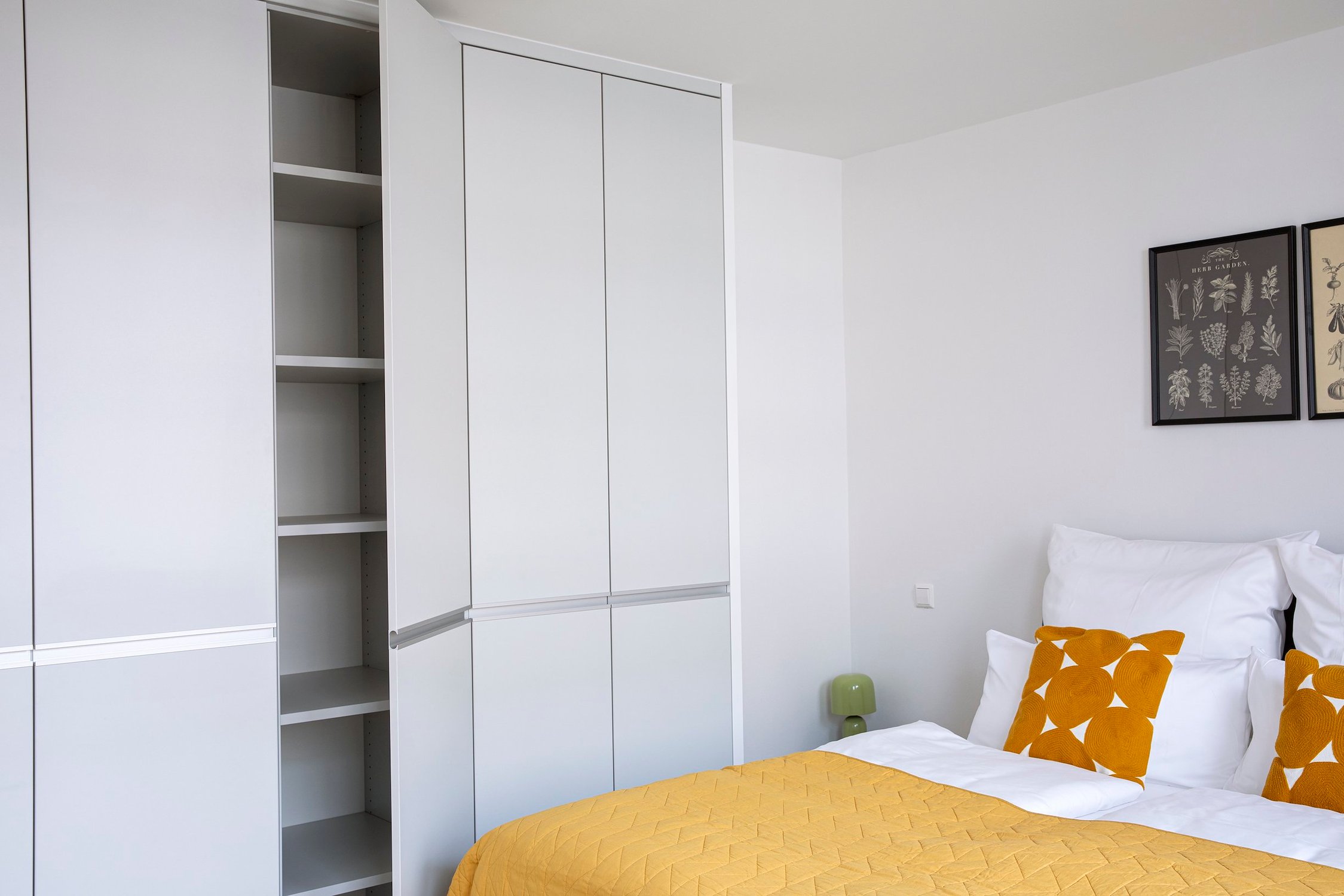 Helles Schlafzimmer mit weißem Einbauschrank, geöffnetem Fachbodenregal und Bett mit gelben Kissen