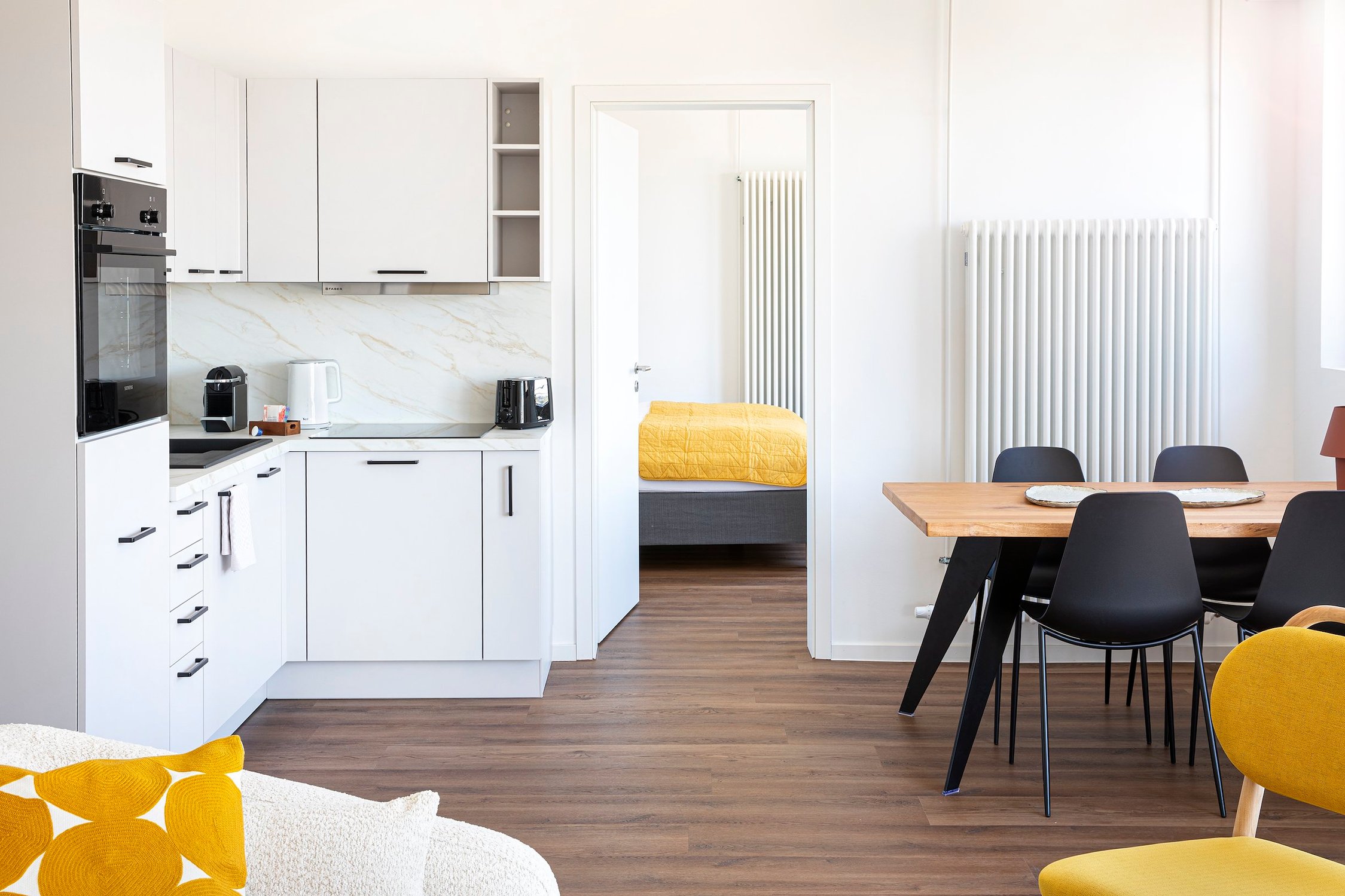 Helle offene Wohnung mit kleiner weißer Küchenzeile, Holztisch, vier schwarzen Stühlen und Heizkörper