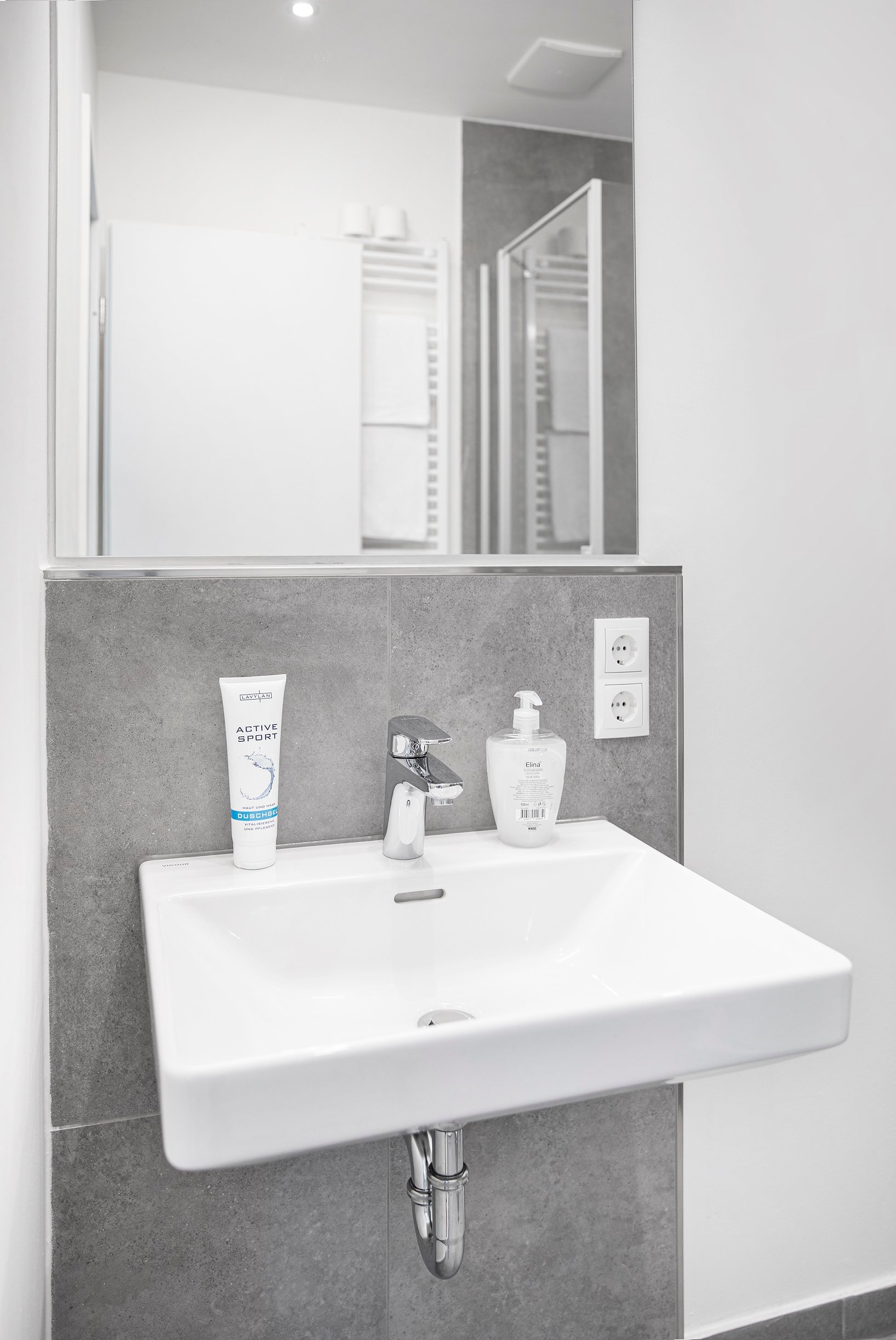 Modernes Badezimmer mit weißem Waschbecken, grauen Fliesen und Spiegel, Duschkabine im Hintergrund