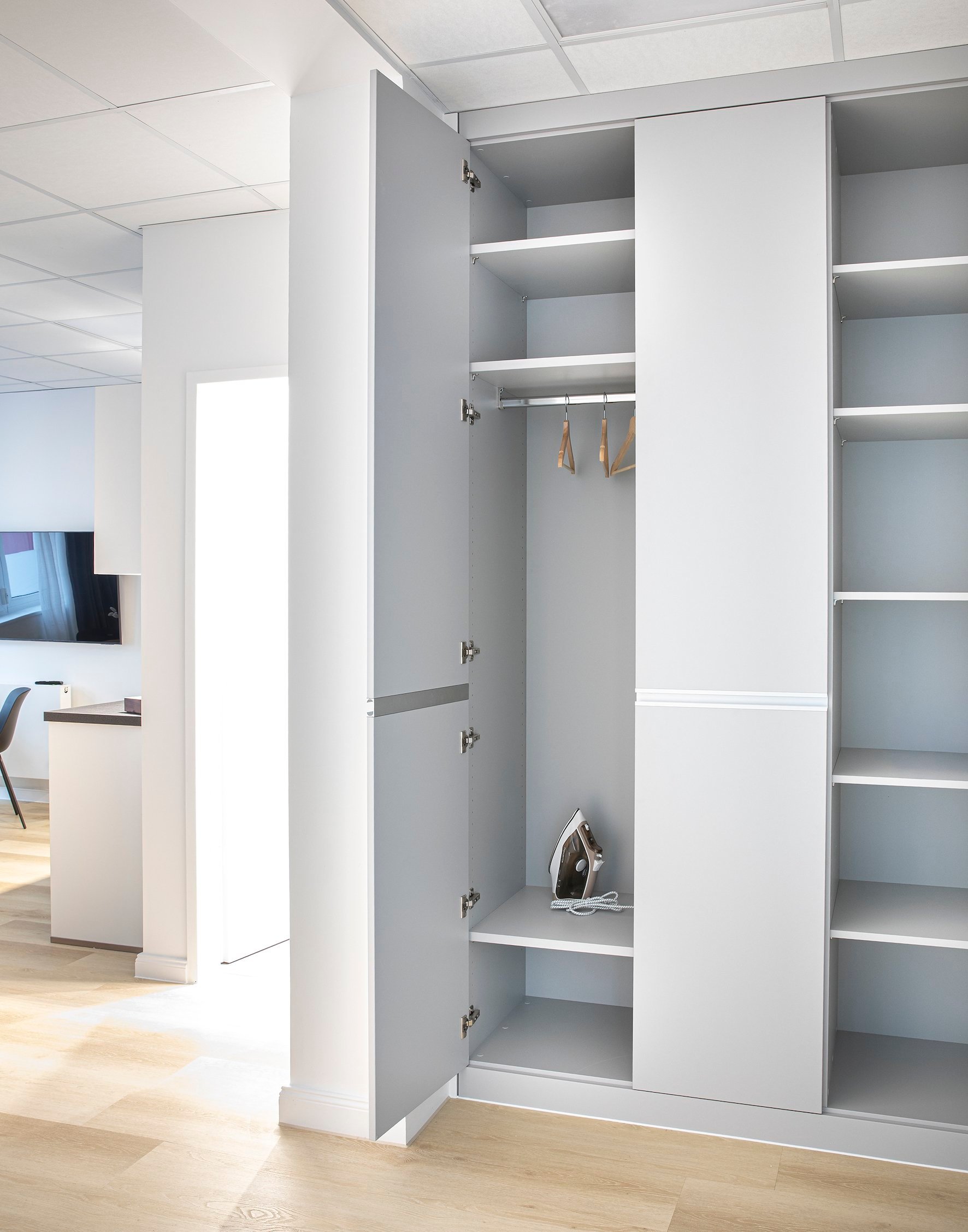 Grauer, offener Kleiderschrank mit Regalen, Kleiderstange und Bügeleisen, heller Holzboden