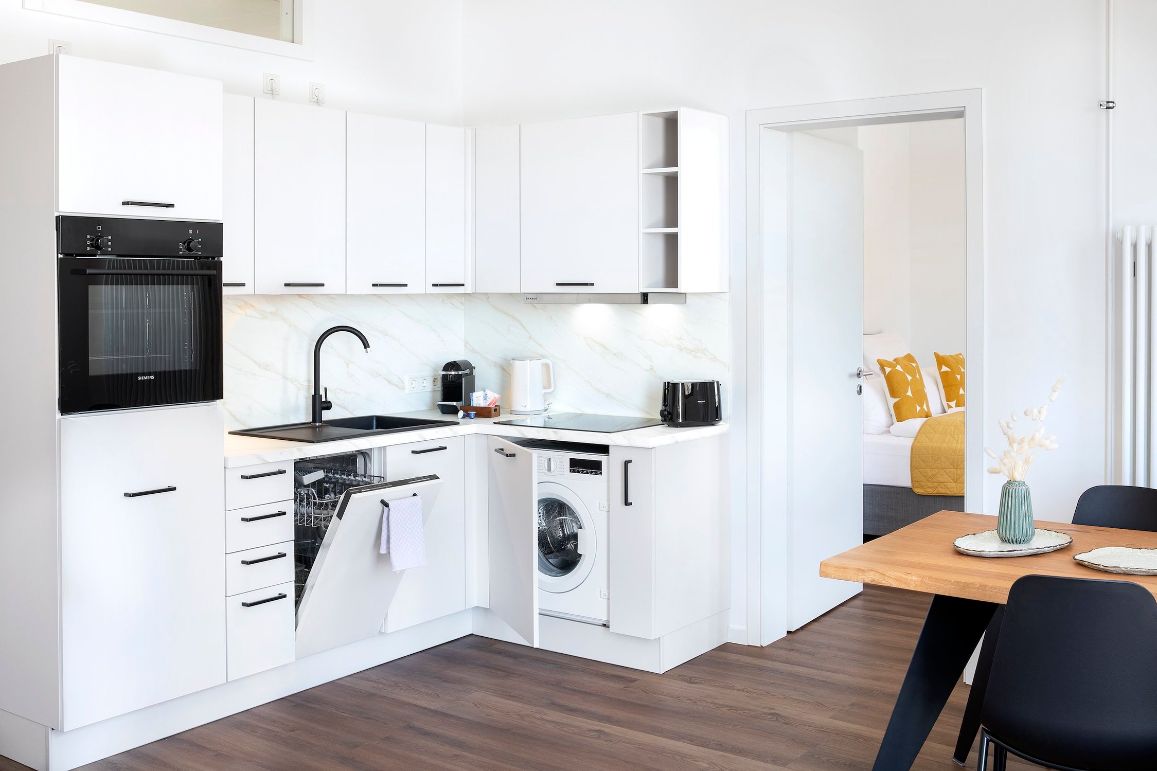 Weiße Einbauküche mit schwarzen Details, Spül- und Waschmaschine geöffnet, Blick ins Schlafzimmer