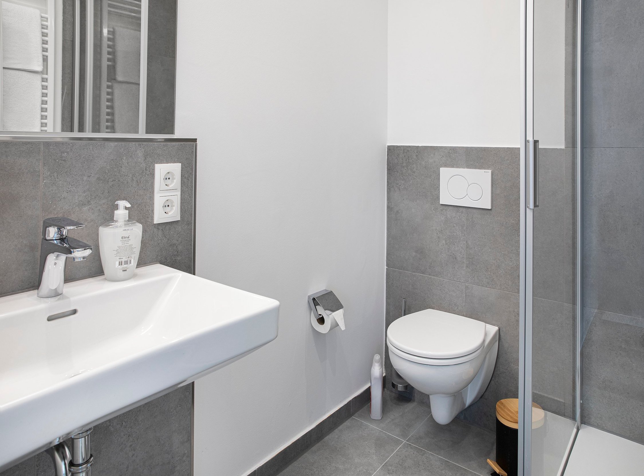 Kleines Badezimmer mit weißem Waschbecken, Toilette rechts und Dusche, graue Fliesen