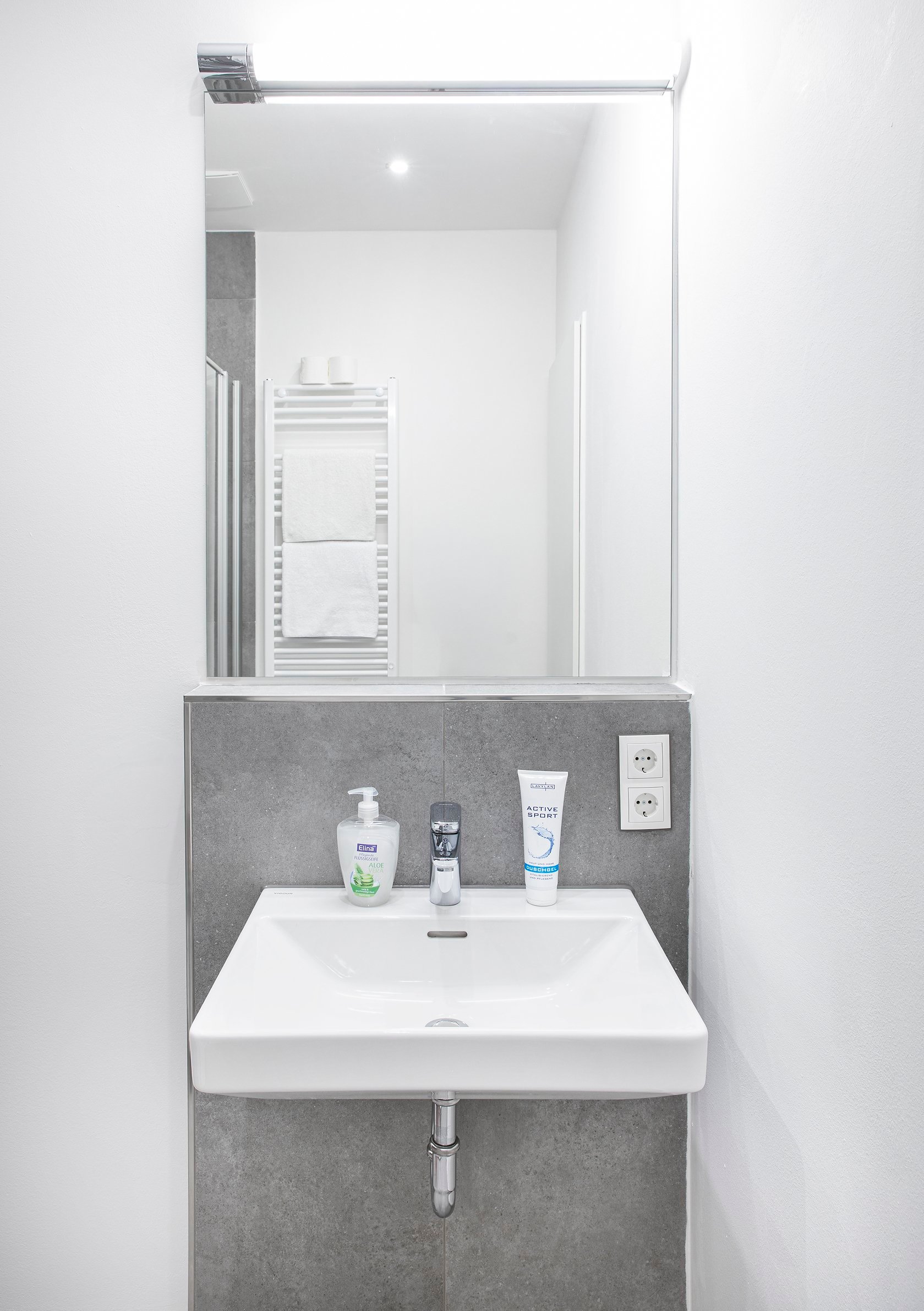 Badezimmer mit weißem Waschbecken, grau gefliester Wand, großem Spiegel und Hygieneartikeln