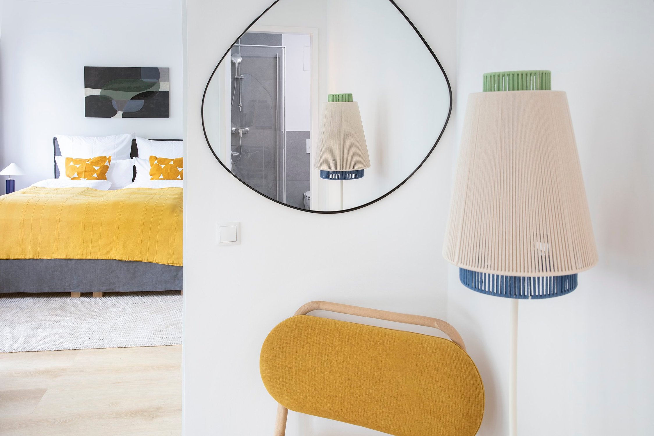 Schlafzimmer mit gelber Bettdecke, abstraktem Bild, daneben Spiegel, Lampe und Hocker