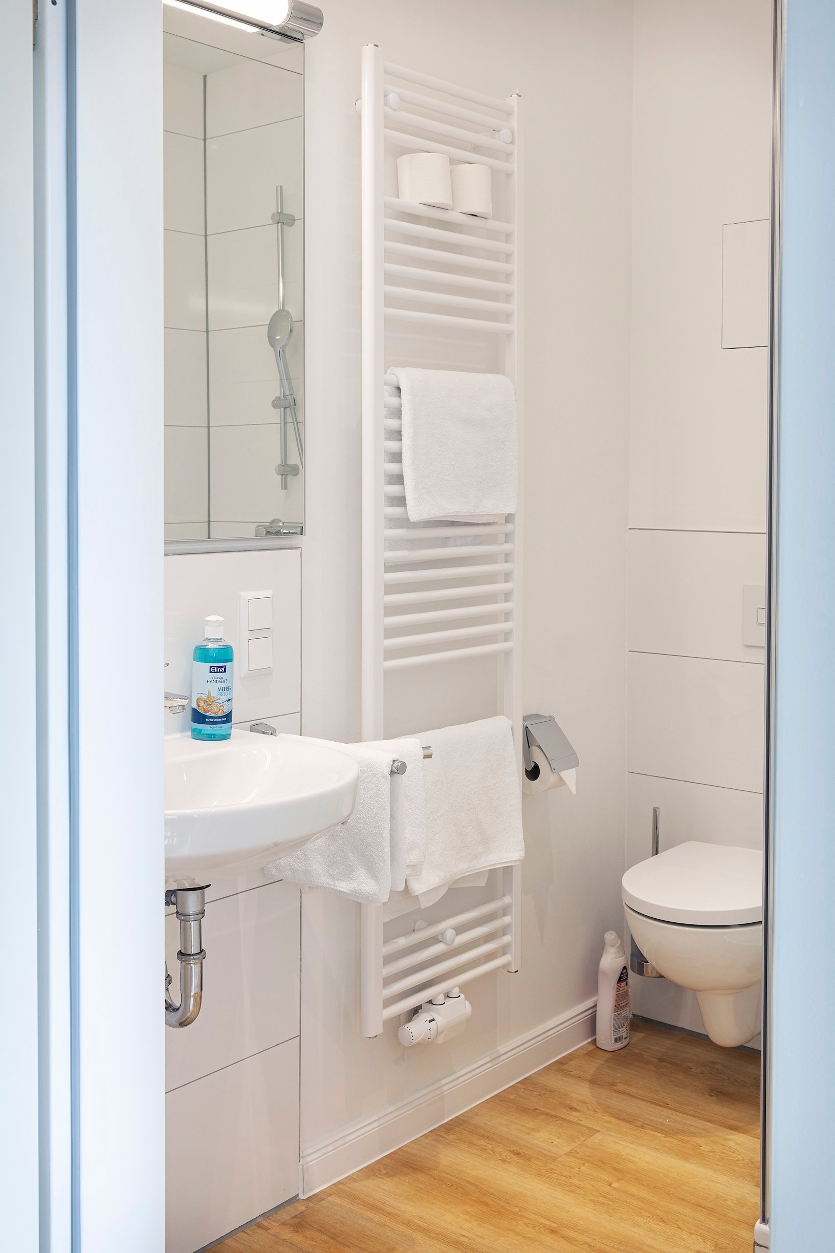Weißes Badezimmer mit Waschbecken, Toilette und Handtuchheizkörper, hellem Holzboden