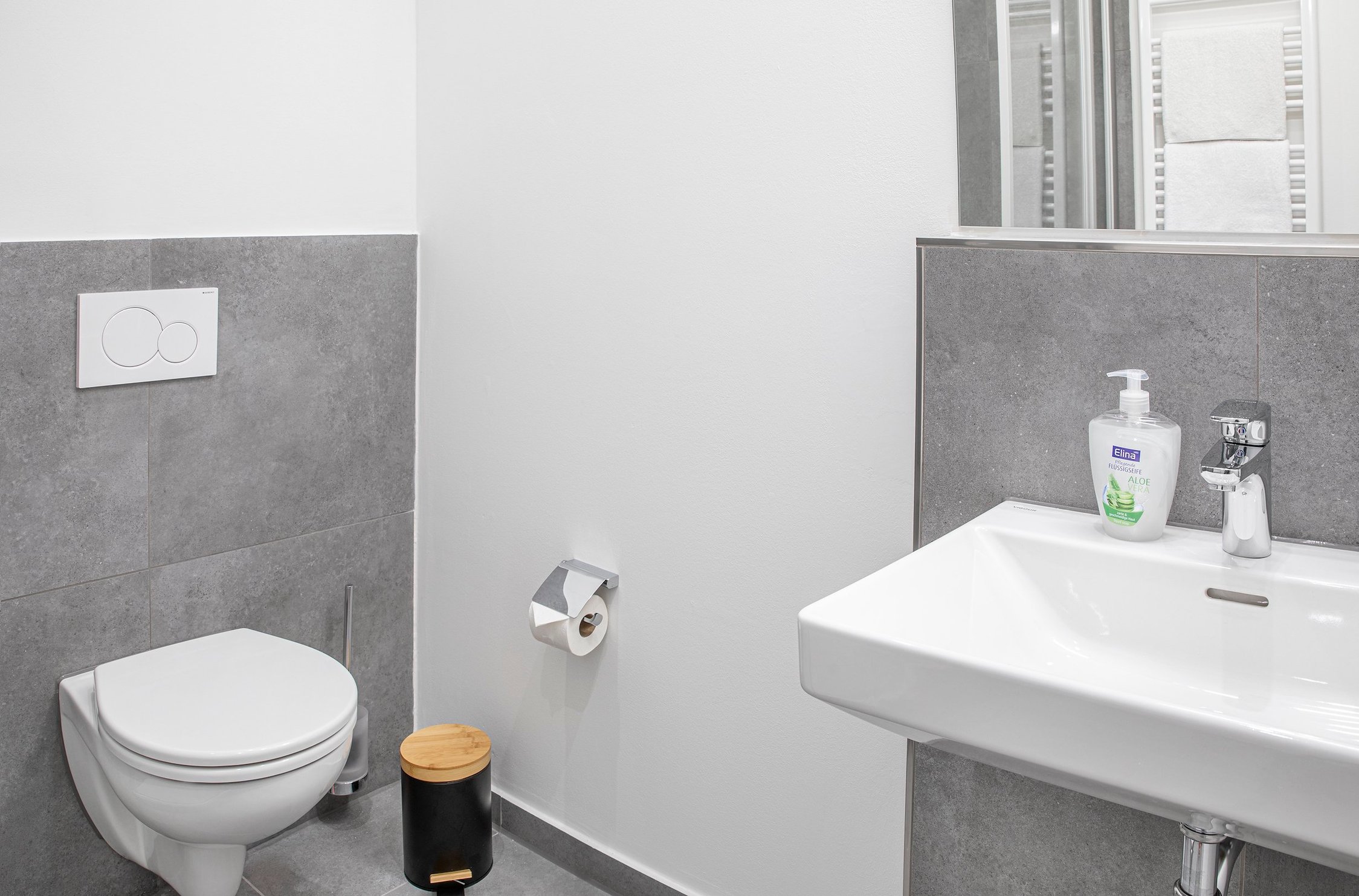 Bad mit weißer Toilette, grau gefliesten Wänden, weißem Waschbecken und Spiegel