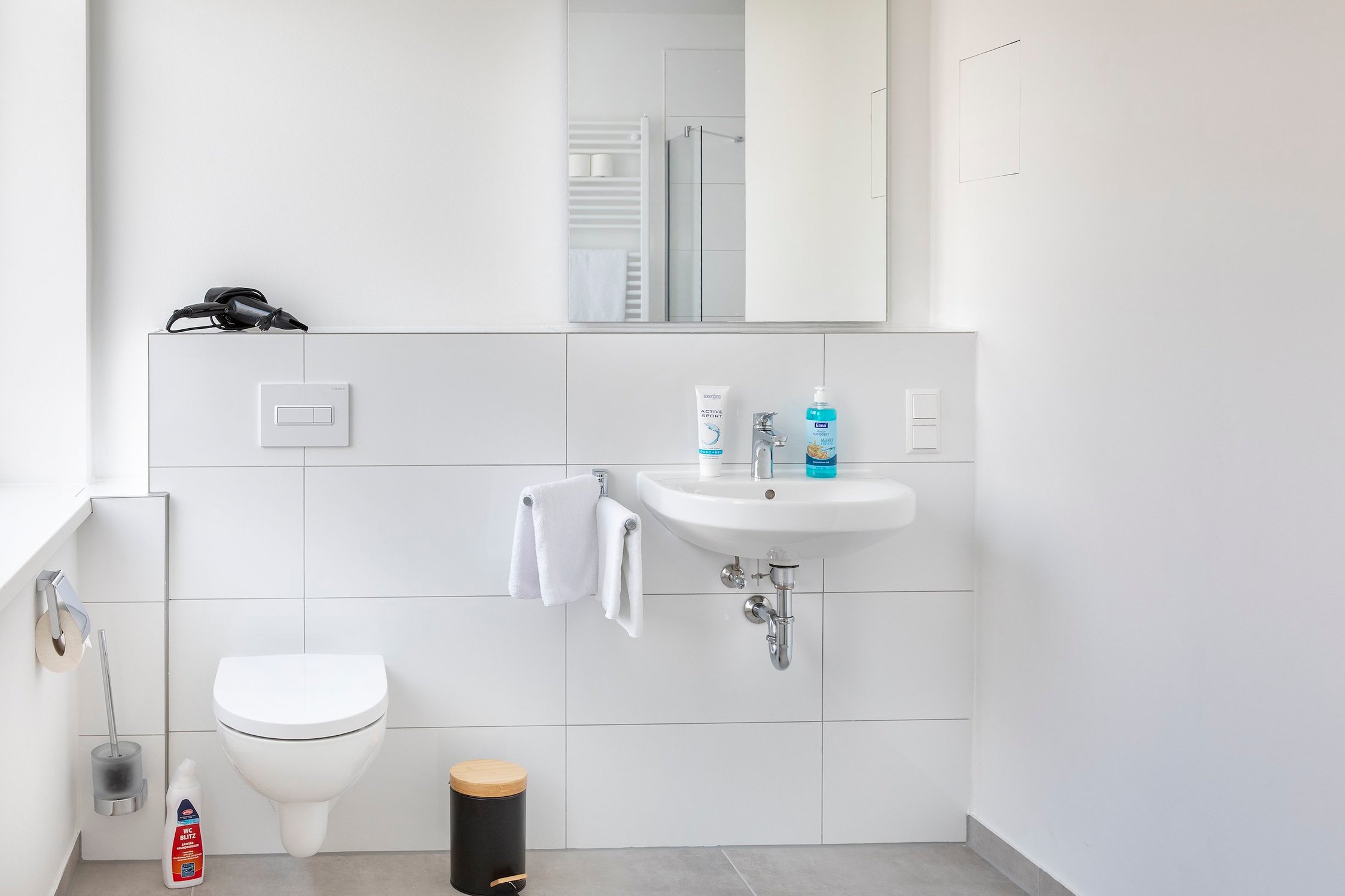 Badezimmer in Weiß mit WC, Waschbecken und Spiegel, dazu schwarzer Mülleimer