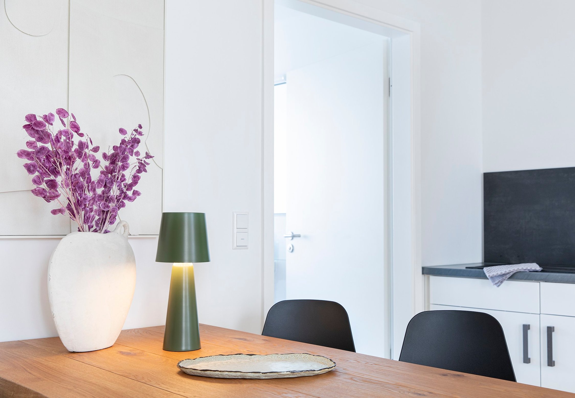 Holztisch mit Vase und lila Blüten, grüner Lampe, Stühlen, Küche und Tür im Hintergrund