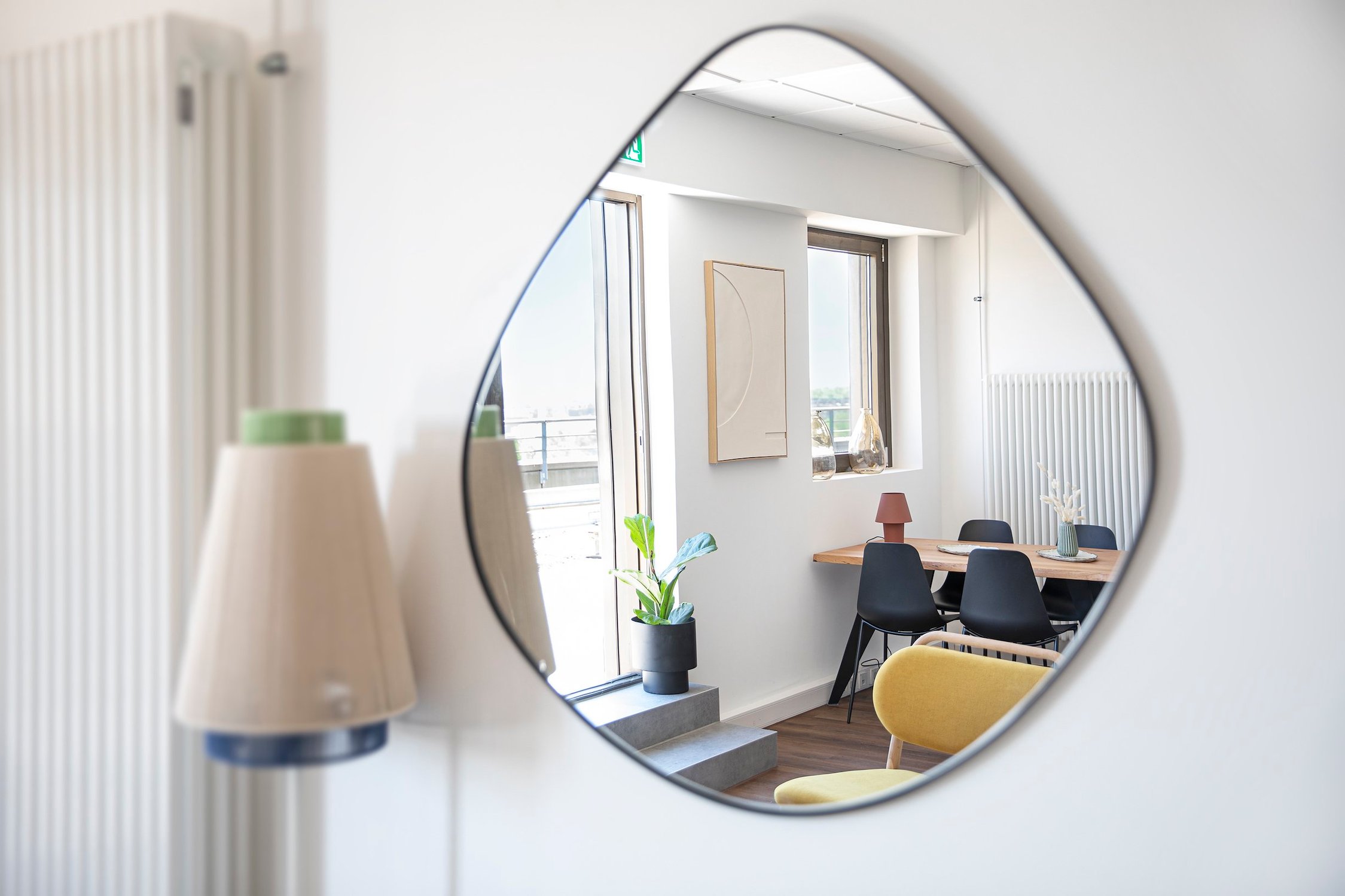 Geschwungener Spiegel an weißer Wand, reflektiert hellen Essbereich mit Holztisch, schwarze Stühle