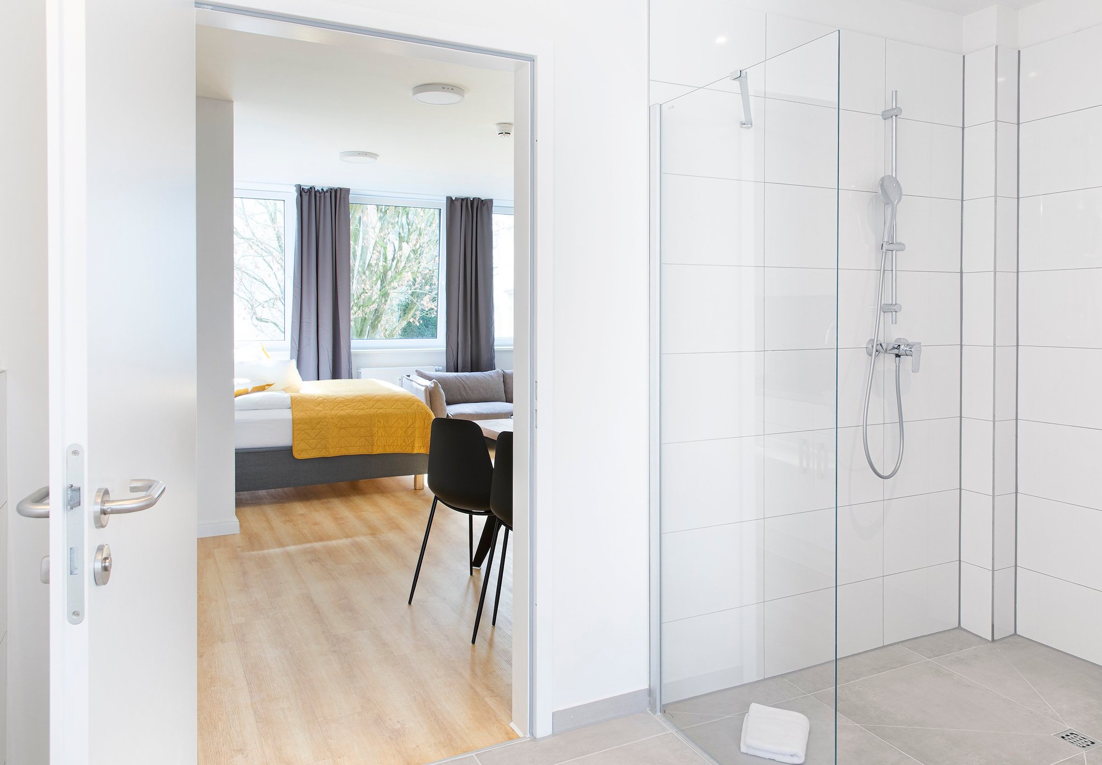 Badezimmer mit bodengleicher Dusche, Türöffnung zu Zimmer mit Bett und Tisch