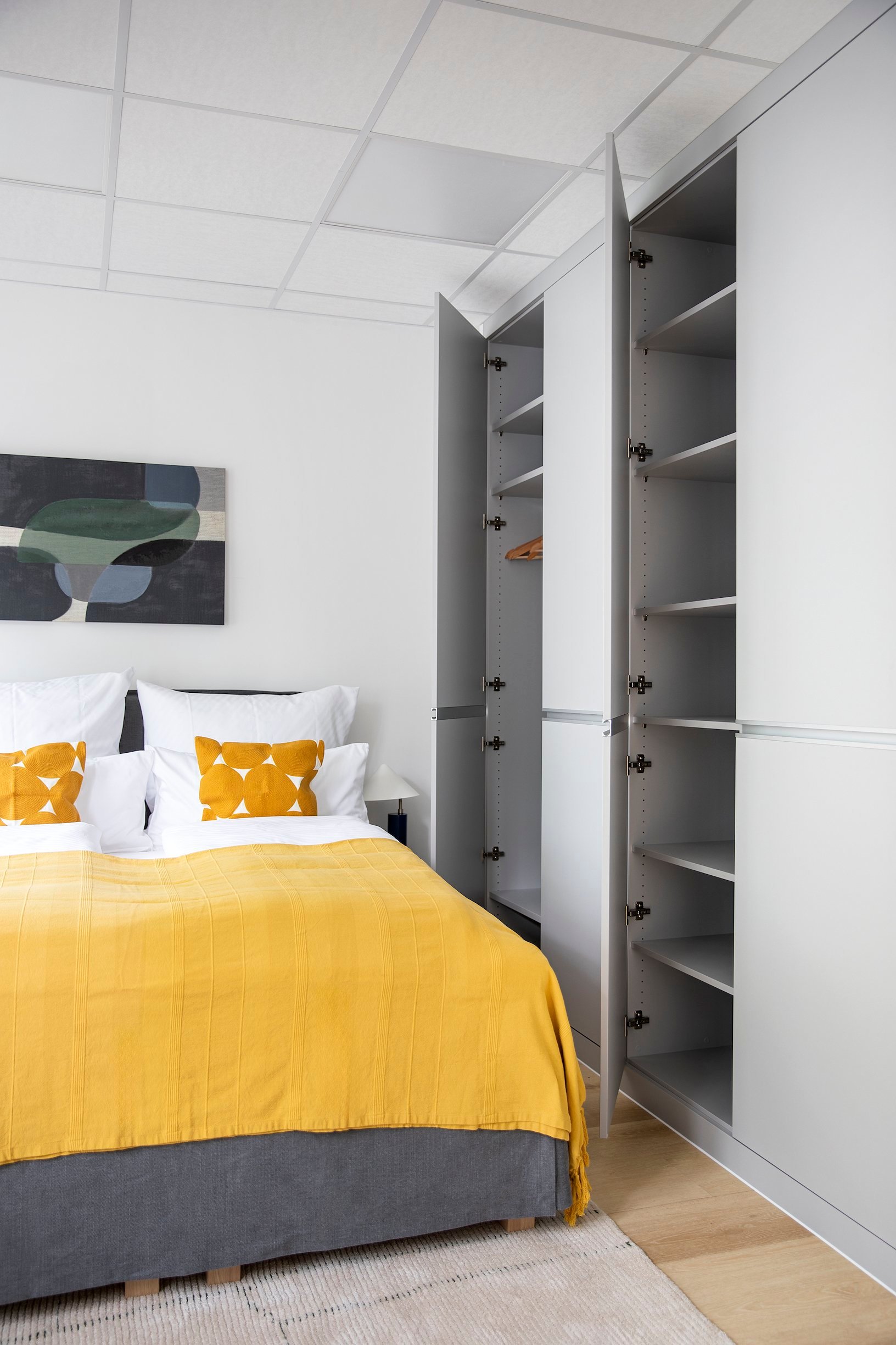 Schlafzimmer mit weißer Bettwäsche, gelber Decke und offenem, grauem Schrank mit Regalen