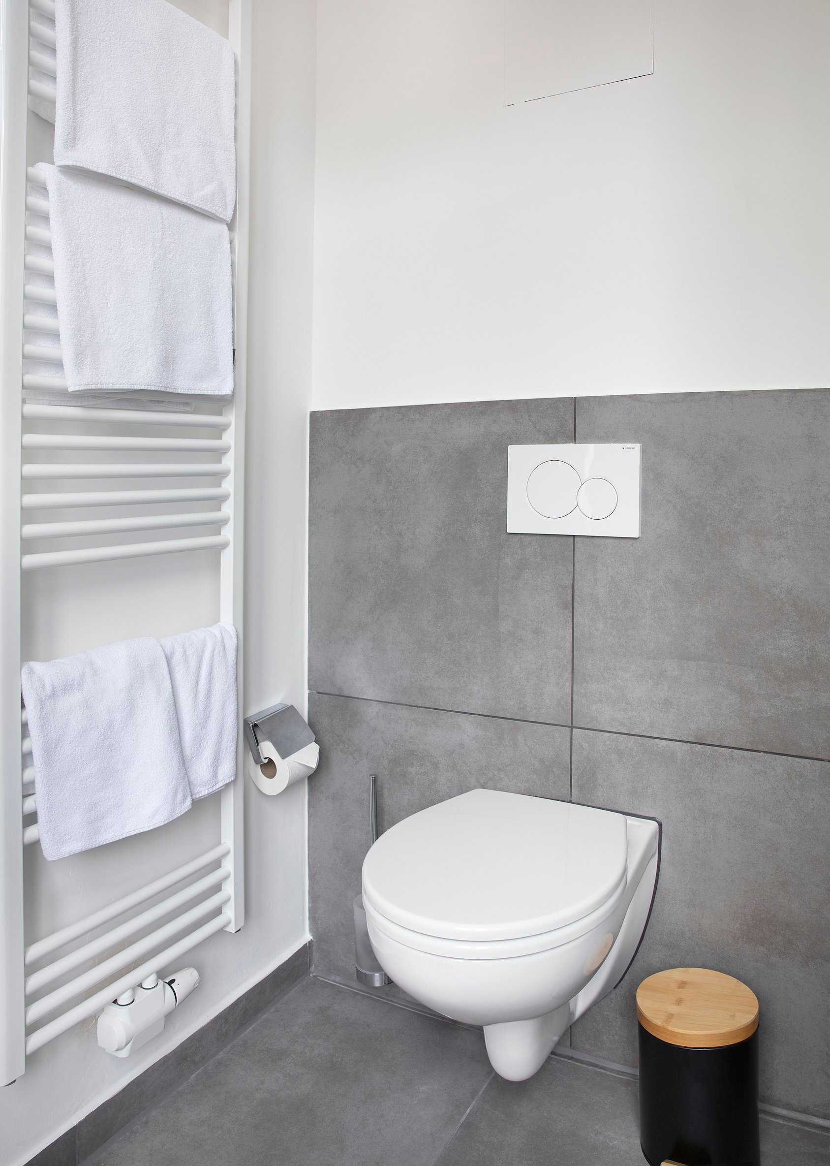 Badezimmer mit weißer Hänge-Toilette, grau gefliesten Wänden und Handtuchheizkörper, hell beleuchtet