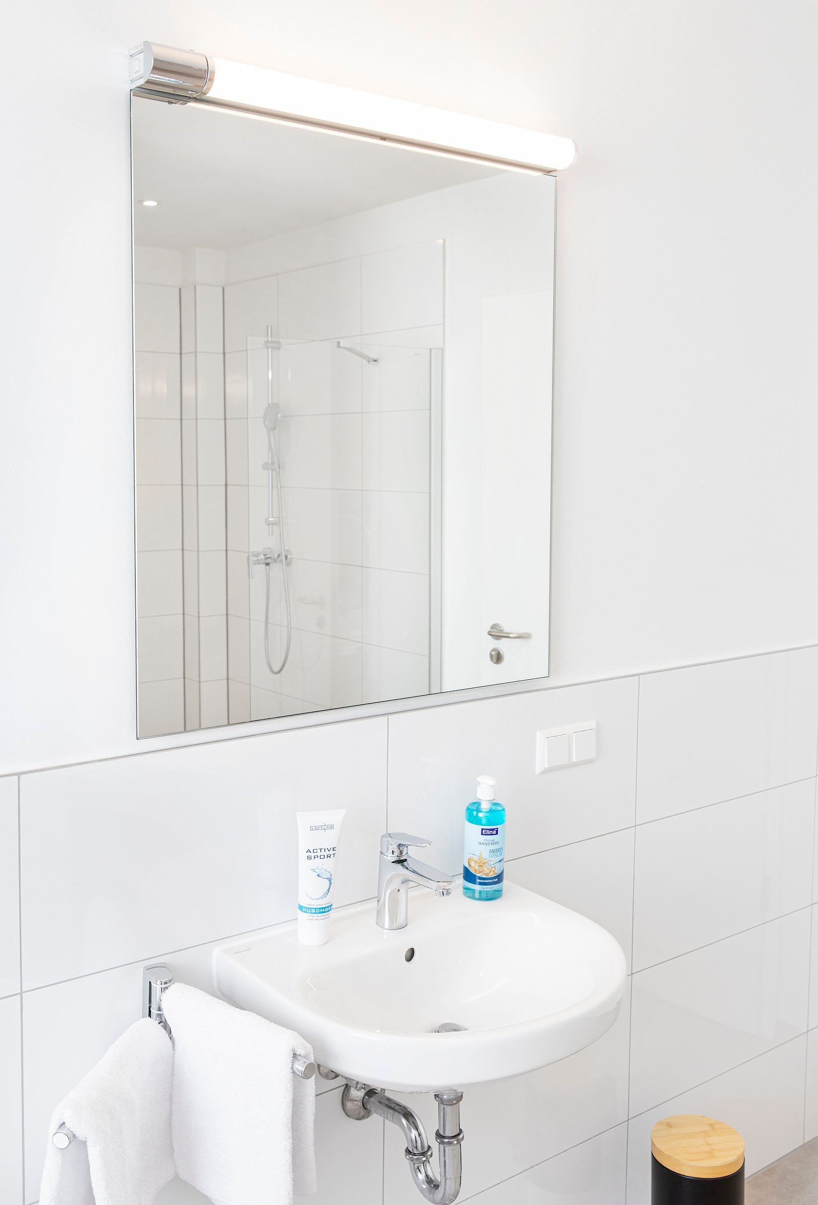 Badezimmer mit weißem Waschbecken, rechteckigem Spiegel, Duschkabine im Spiegel reflektiert