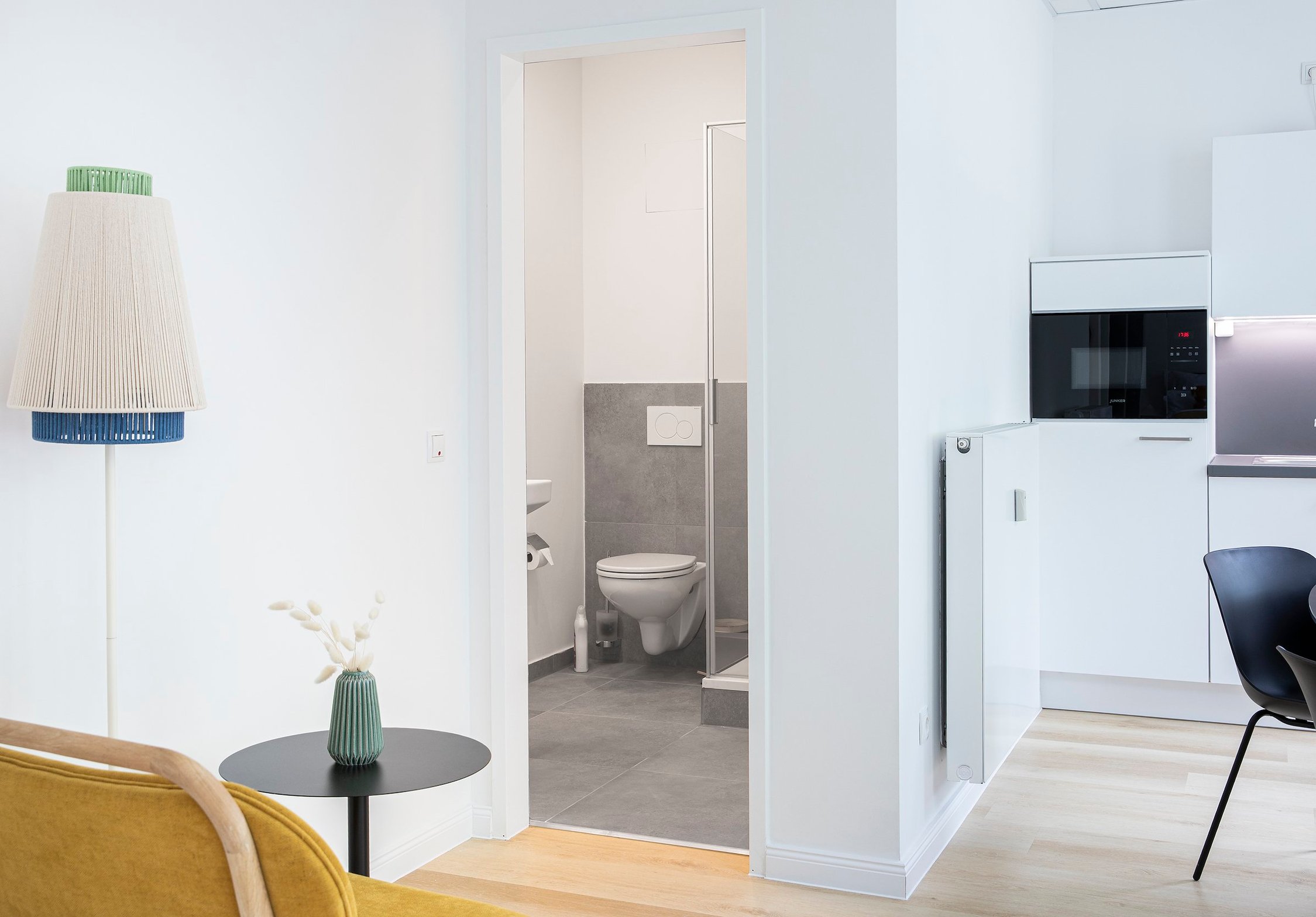 Offener Wohnbereich mit gelbem Sessel, schwarzem Tisch, blau-grüner Wandleuchte, Badezimmer und Küche