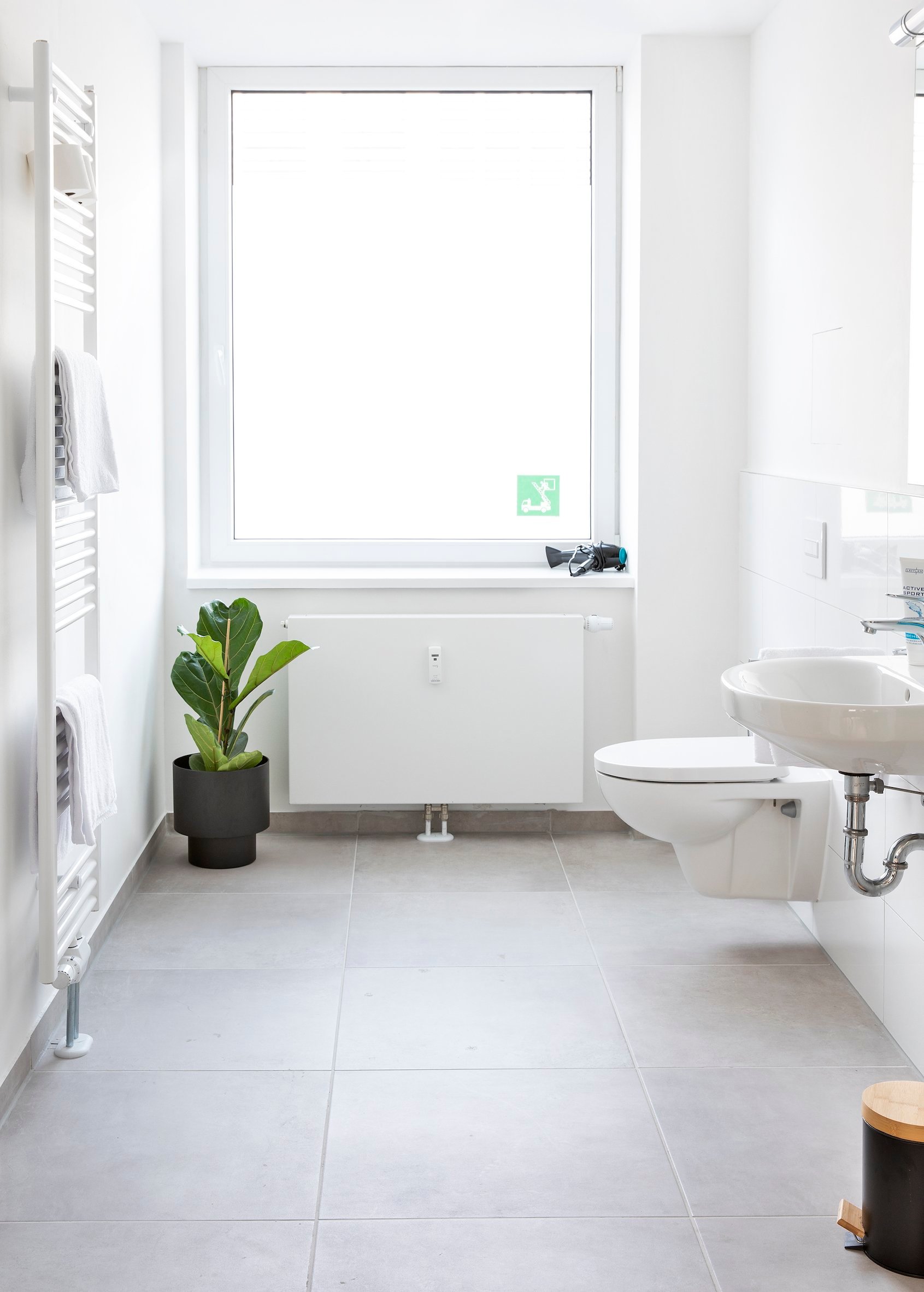 Helles Badezimmer mit weißen Fliesen, Toilette, Waschbecken und Pflanzen vor großem Fenster