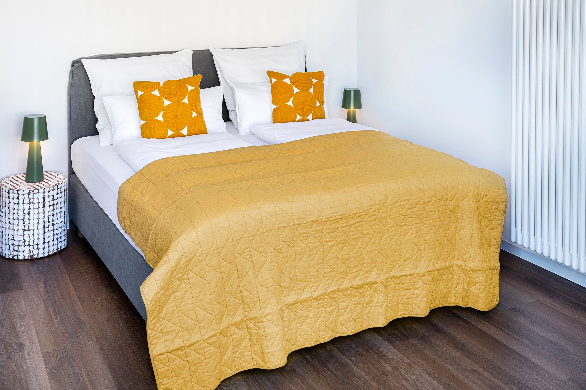 Doppelbett mit gelber Decke und Kissen, dunklem Holzboden, zwei grünen Tischlampen