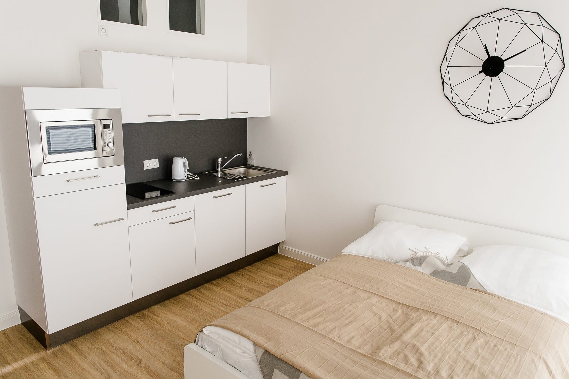 Helles Zimmer mit kleiner Küchenzeile am hellen Holzboden, weißes Bett an weißer Wand