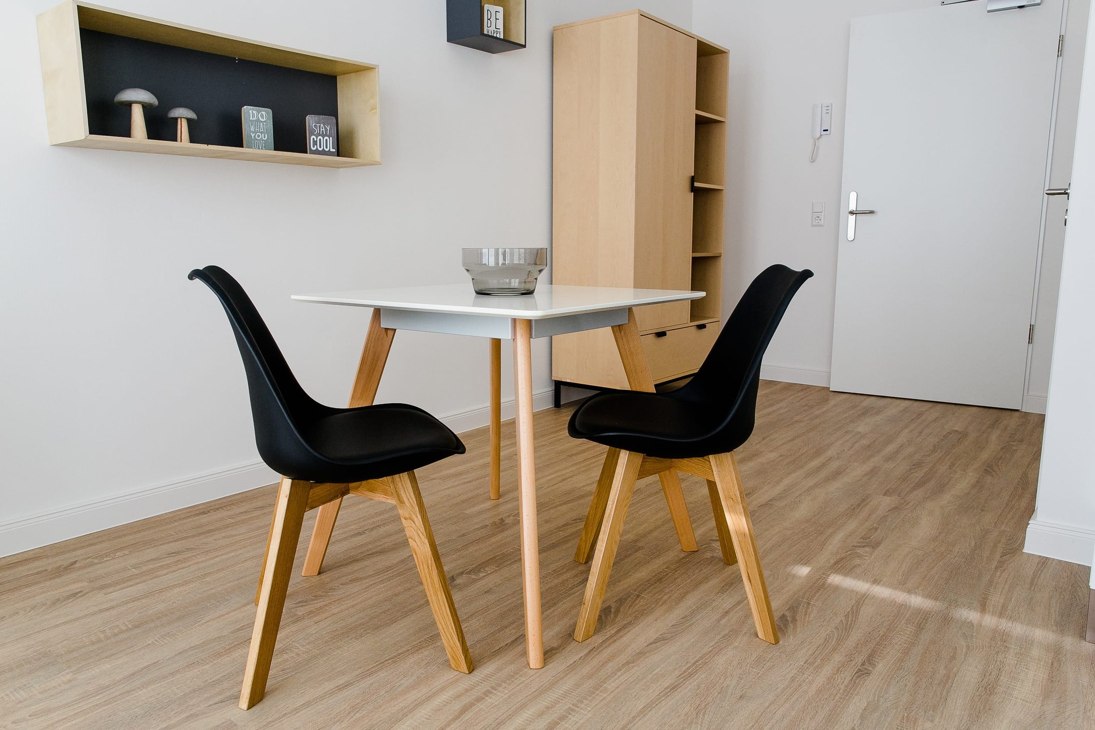 Wohnraum mit hellem Holzboden, weißem Tisch und zwei schwarzen Designerstühlen, Holzregal
