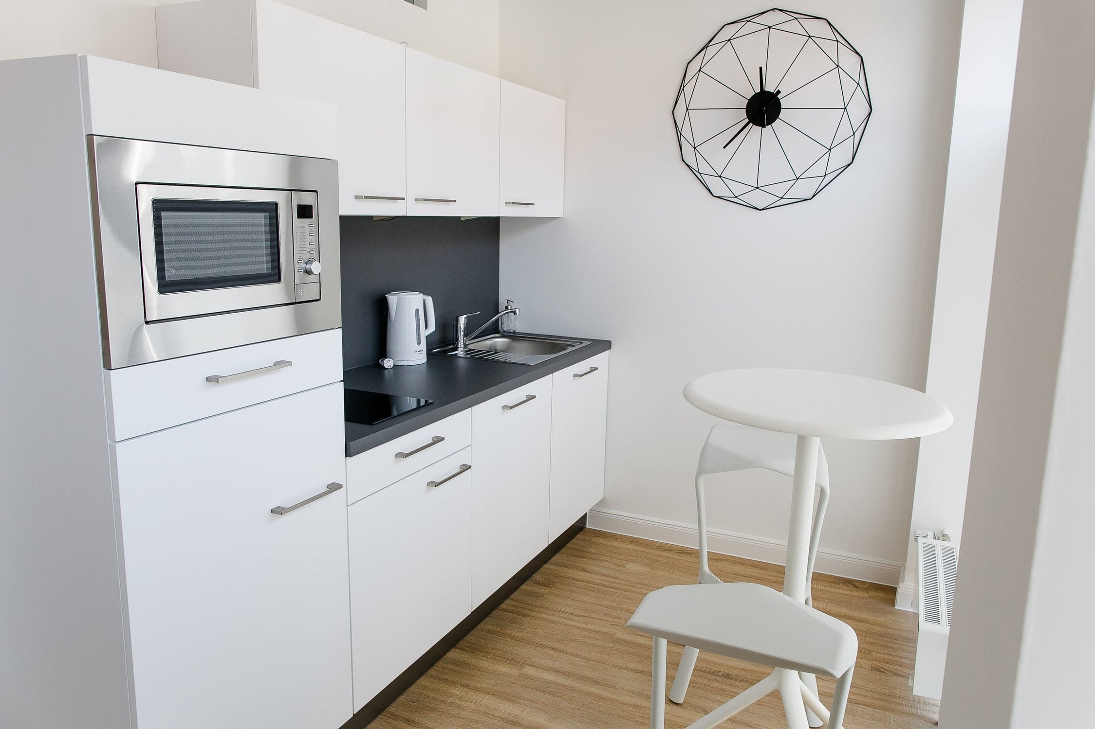 Küche mit weißen Schränken, Mikrowelle und Spüle, dazu runder Tisch und Stuhl