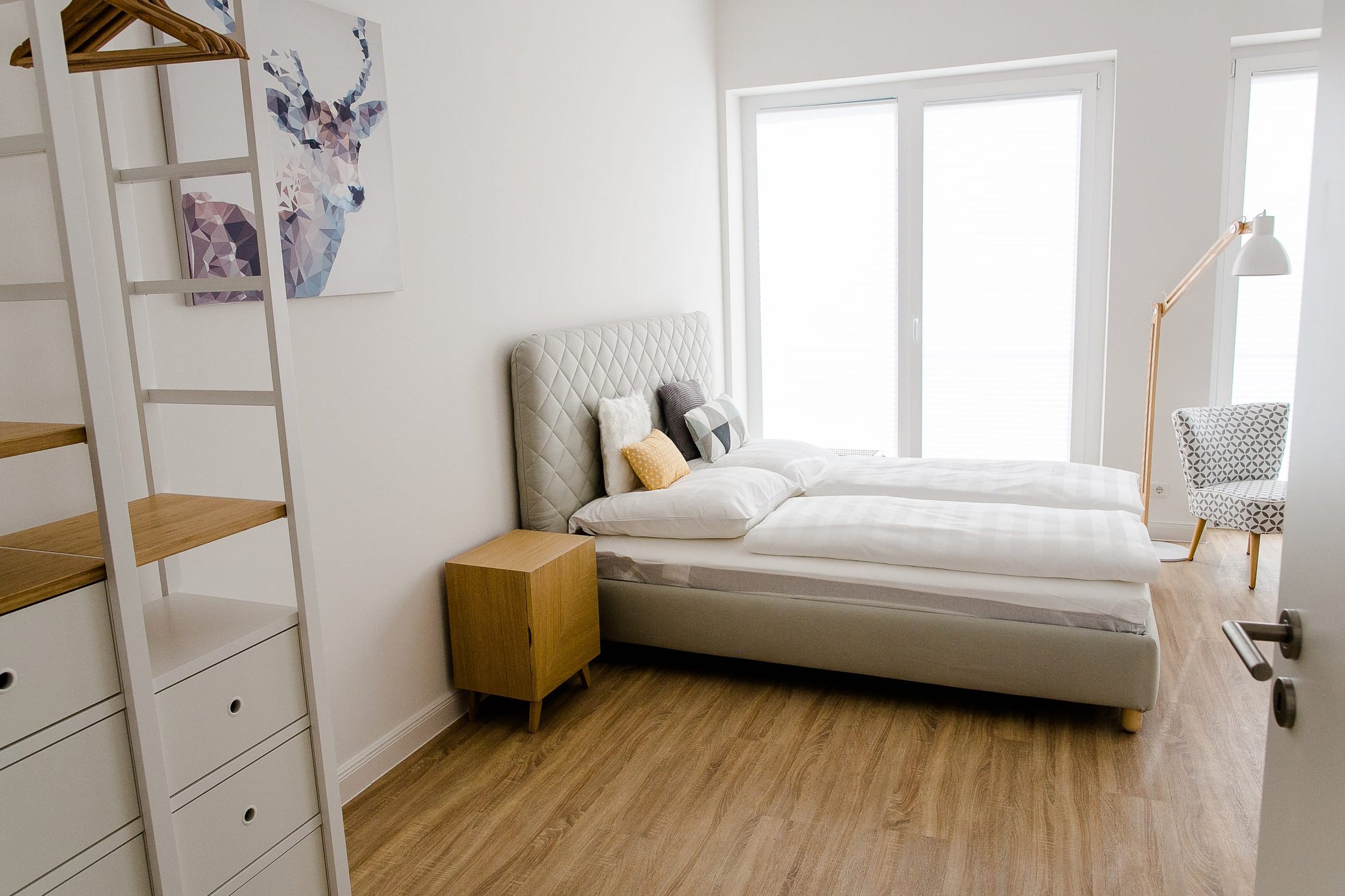 Helles Schlafzimmer mit Holzboden, weißem Bett, Fenster, Stehlampe und Sessel