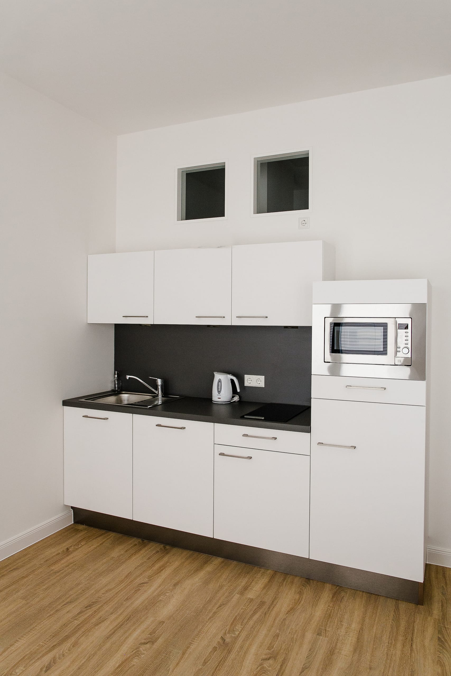 Kleine weiße Küchenzeile mit schwarzer Arbeitsfläche und holzfarbenem Boden in hellem Raum