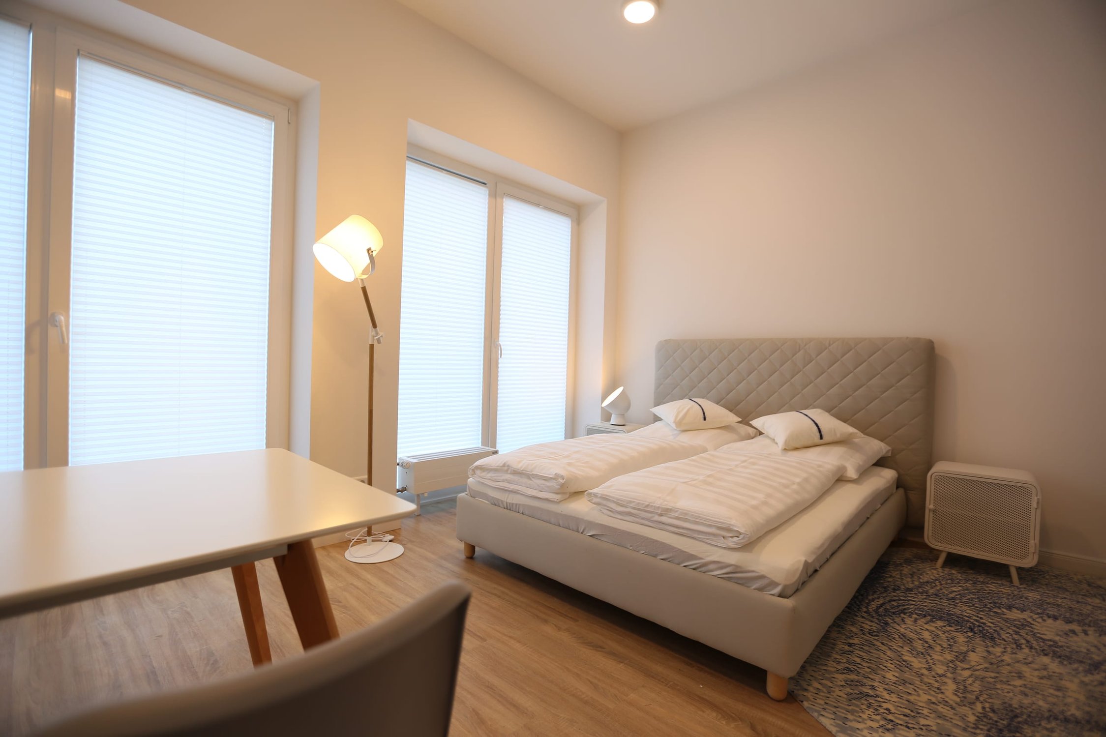 Schlafzimmer mit weißem Bett, zwei Fenstern mit Rollos, Stehlampe und Holzfußboden