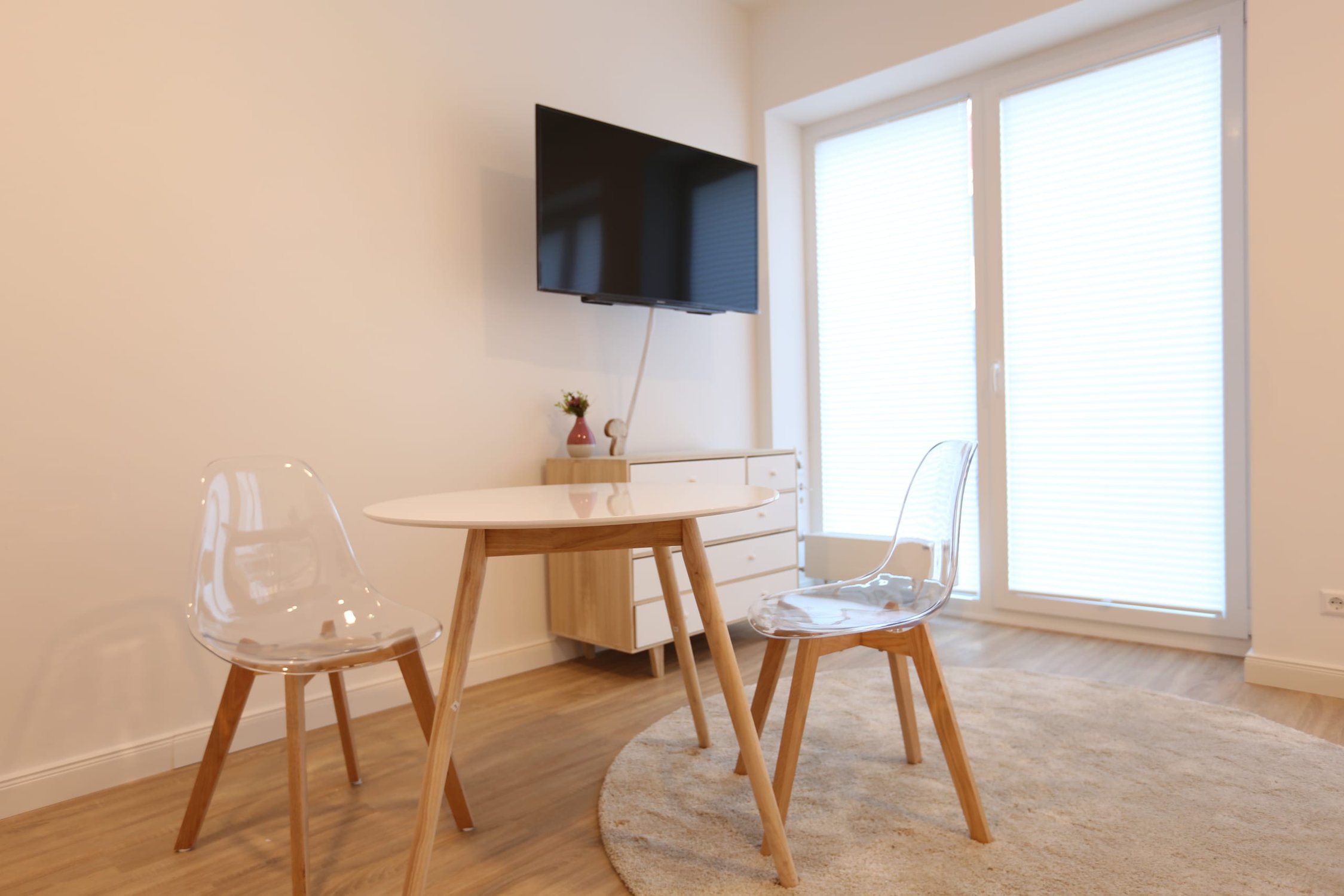 Essbereich mit hellem Holztisch, zwei transparenten Stühlen, Flachbildfernseher an der Wand