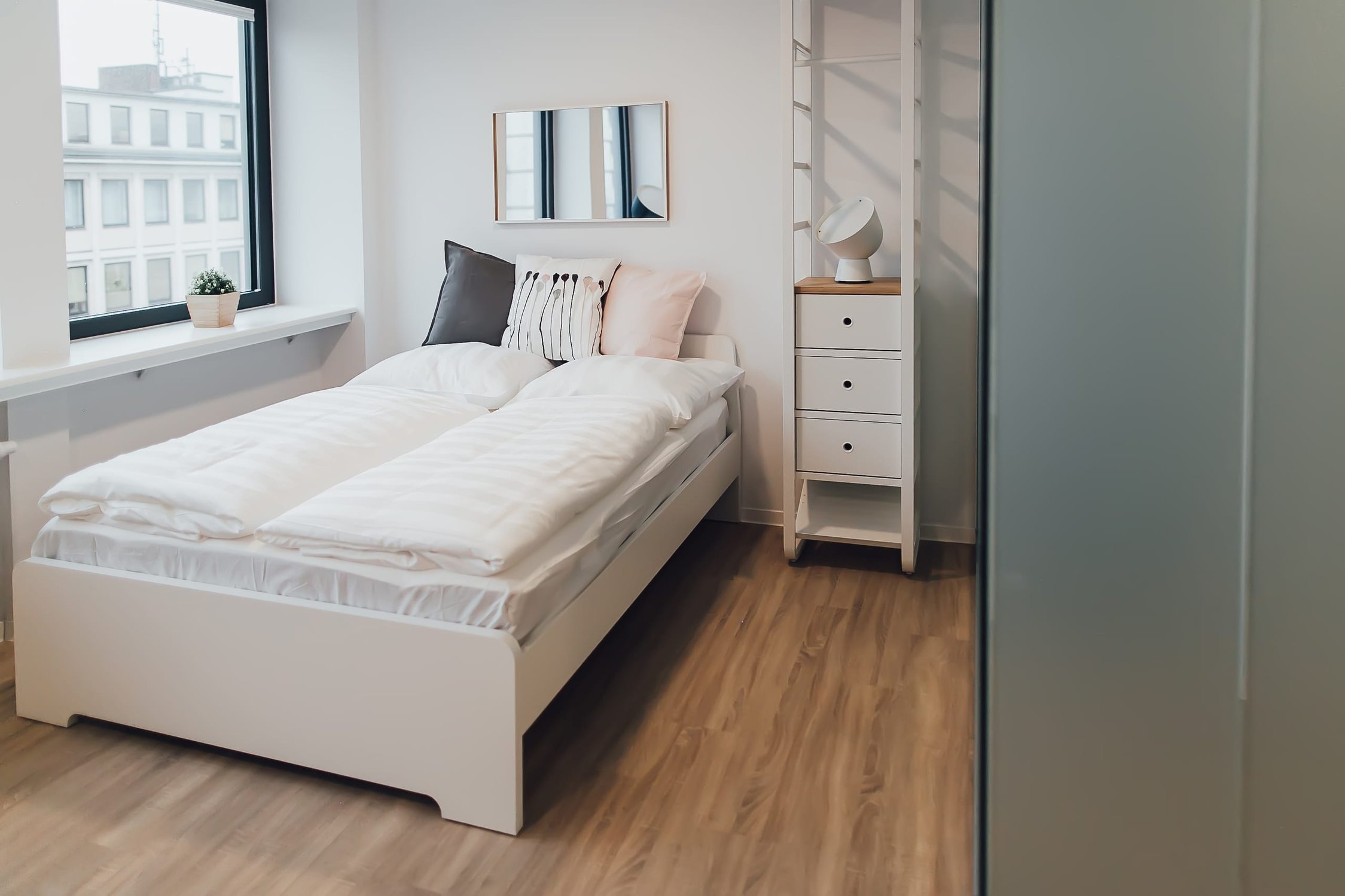 Schlafzimmer mit weißem Bett, Fenster, Holzdielen und Regal mit drei Schubladen