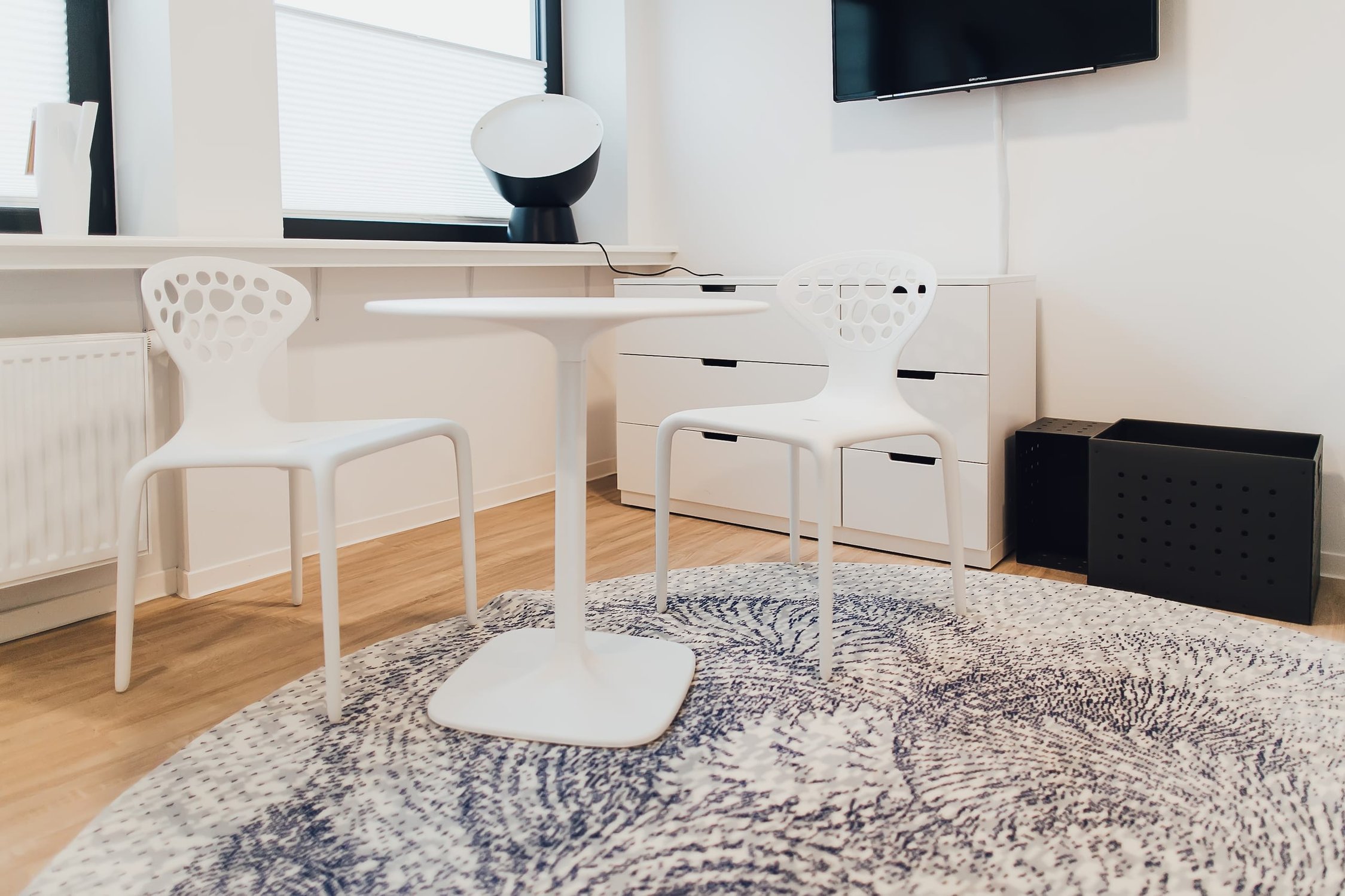 Helles Zimmer mit zwei weißen Stühlen, rundem Tisch, grauem Teppich und Kommode