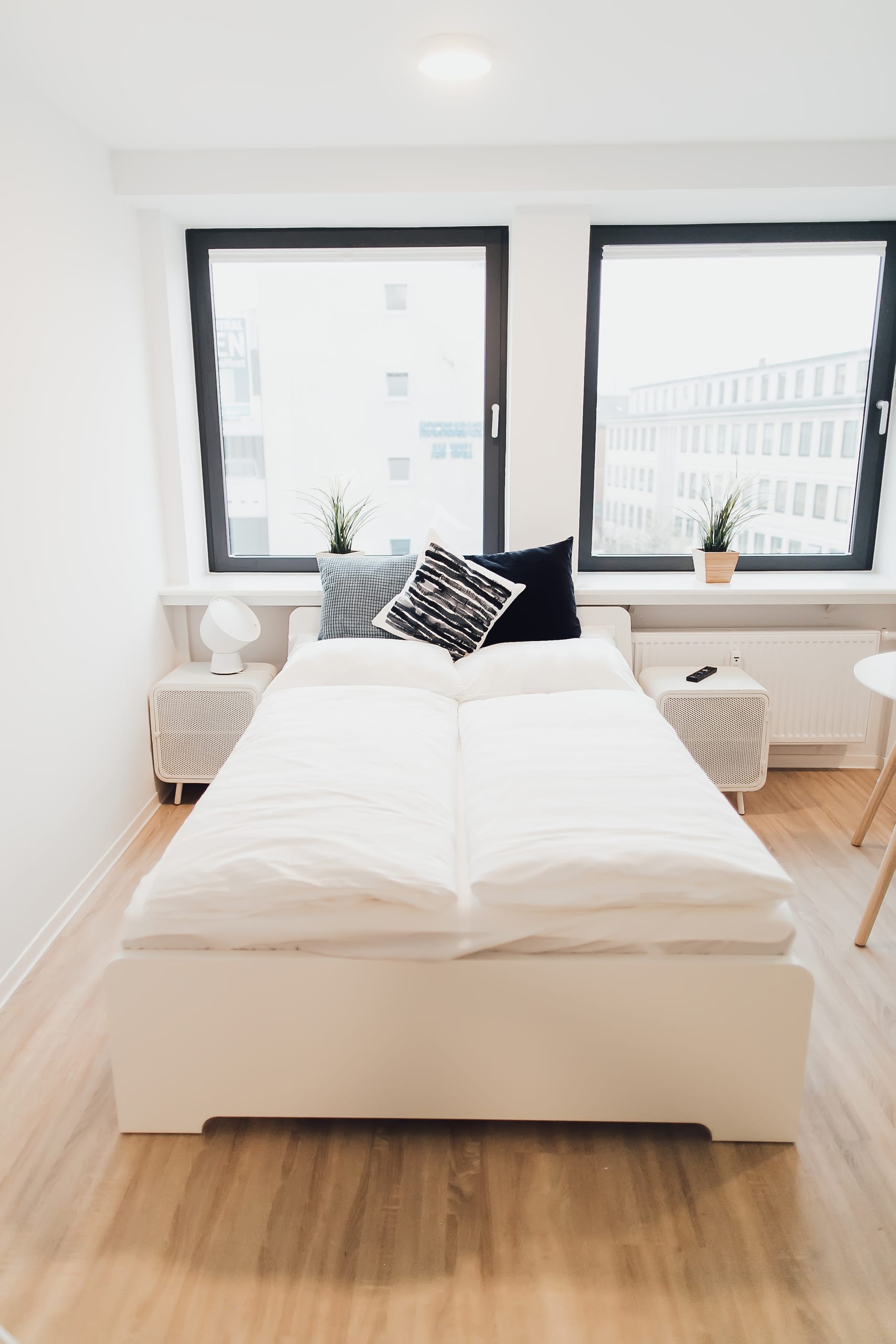 Schlafzimmer mit weißem Bett, holzfarbenem Boden und zwei großen Fenstern