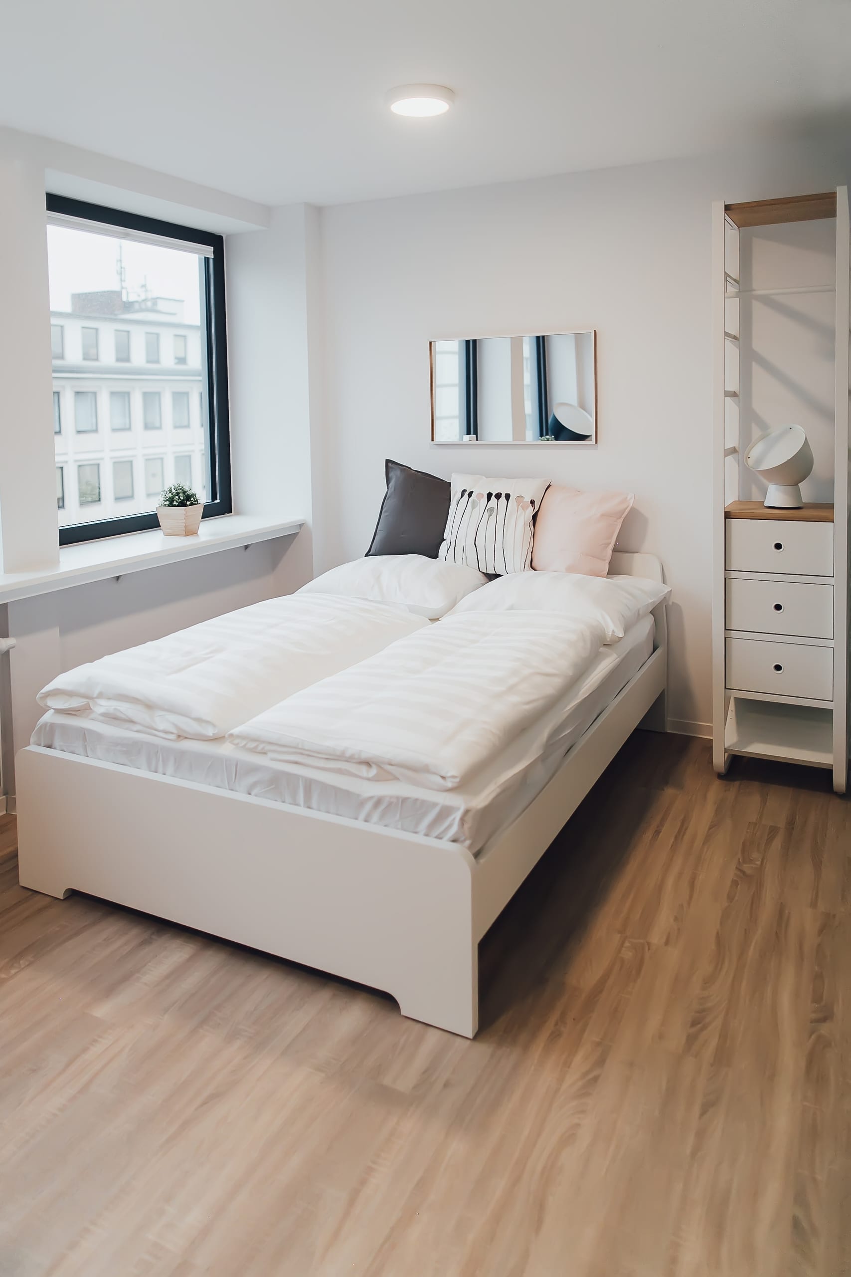 Schlafzimmer mit weißem Doppelbett, Spiegel und hellbraunem Holzboden