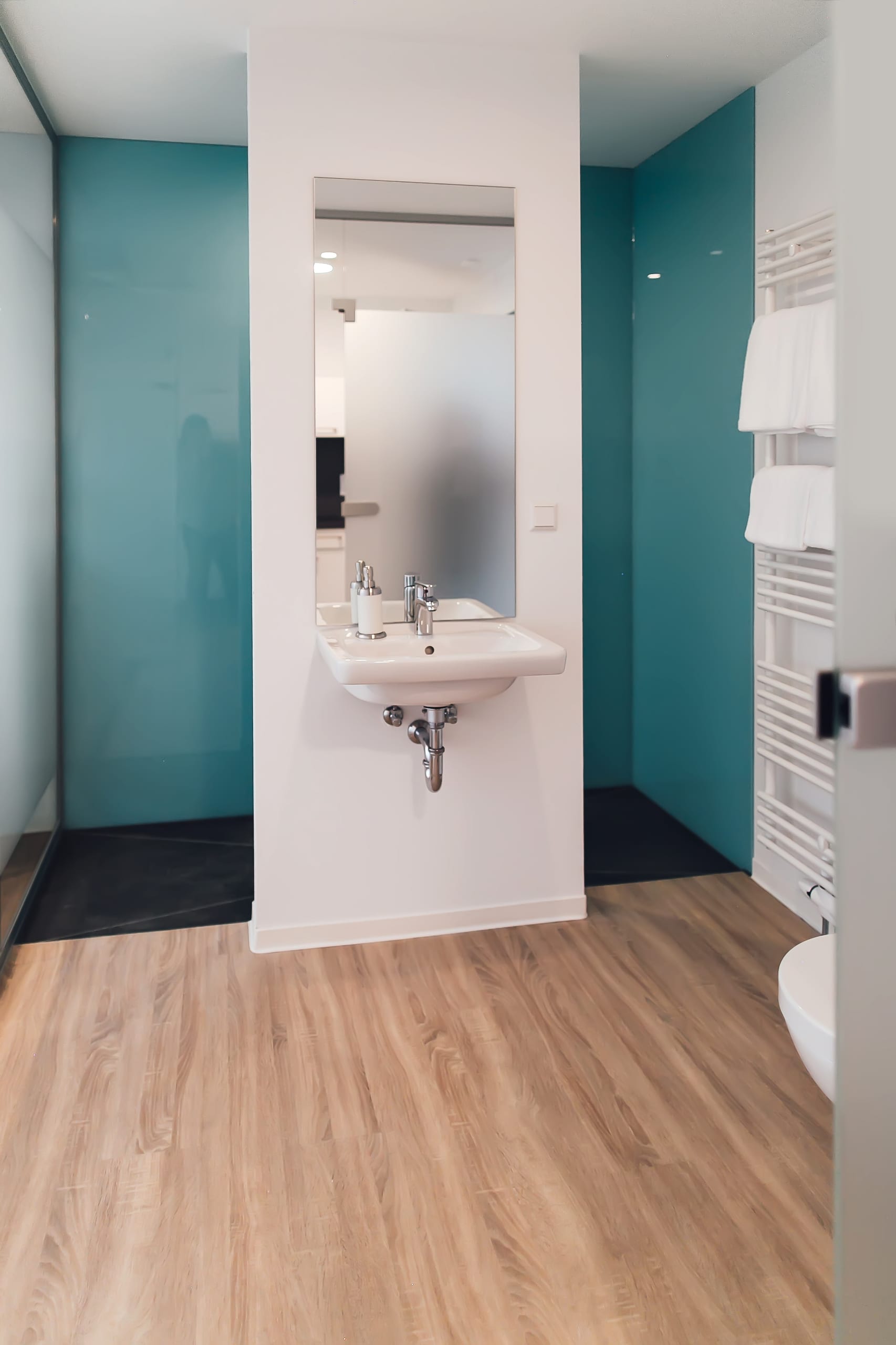 Modernes Badezimmer mit Waschtisch, Spiegel, hellbraunem Holzboden, tealblauen Wänden und Handtuchhalter